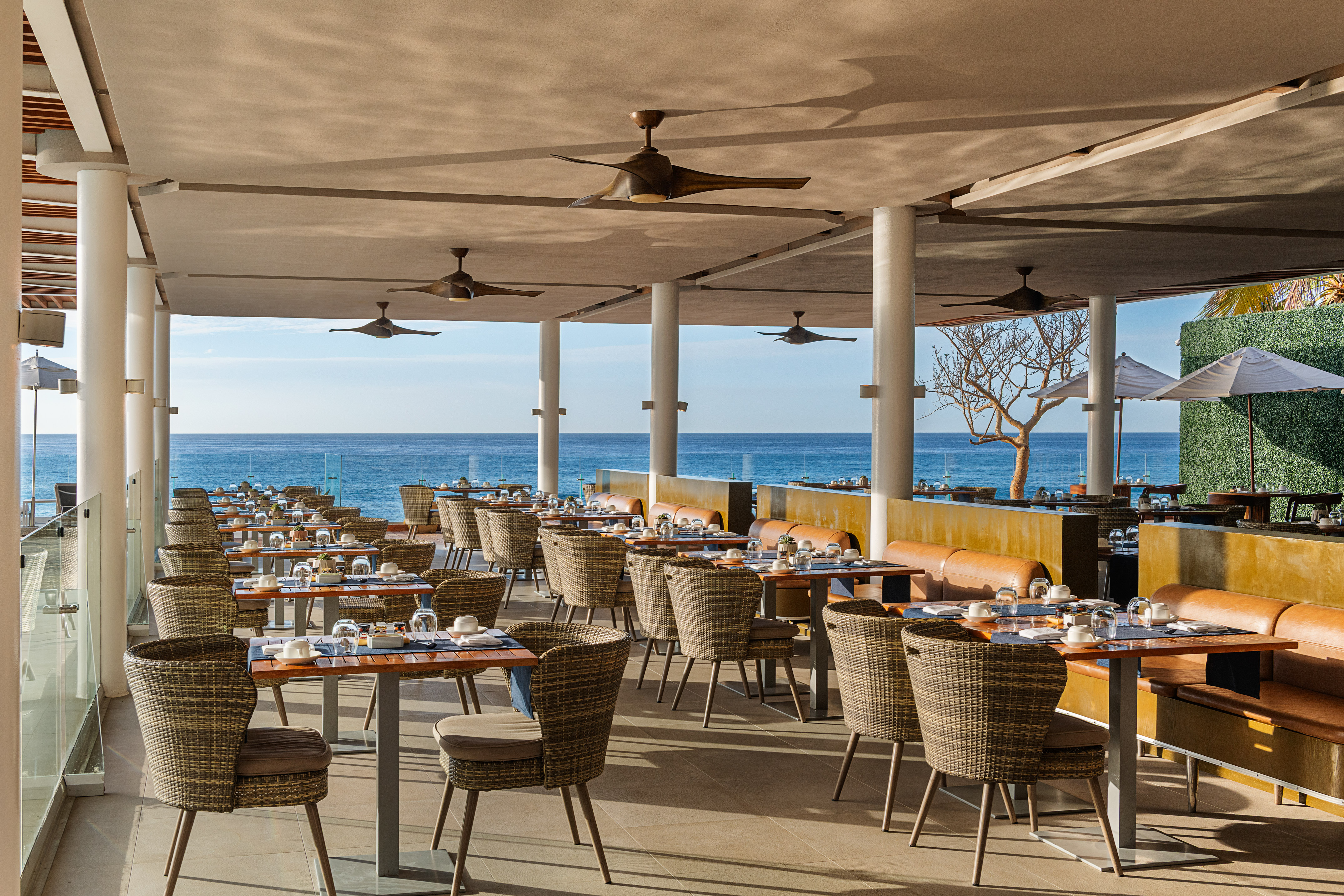 Outdoor restaurant patio with ocean view, set tables, and wicker chairs.