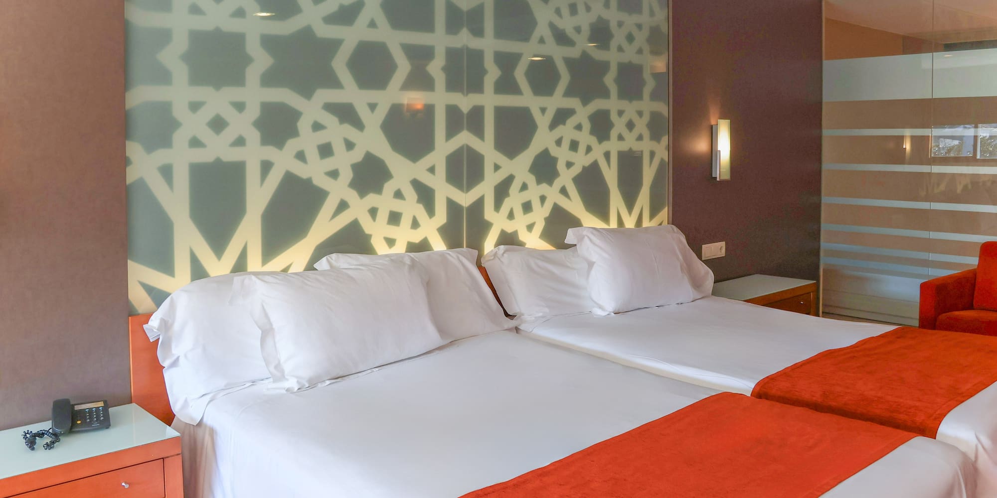 Hotel room with twin beds, white bedding, orange throws, and geometric headboard.