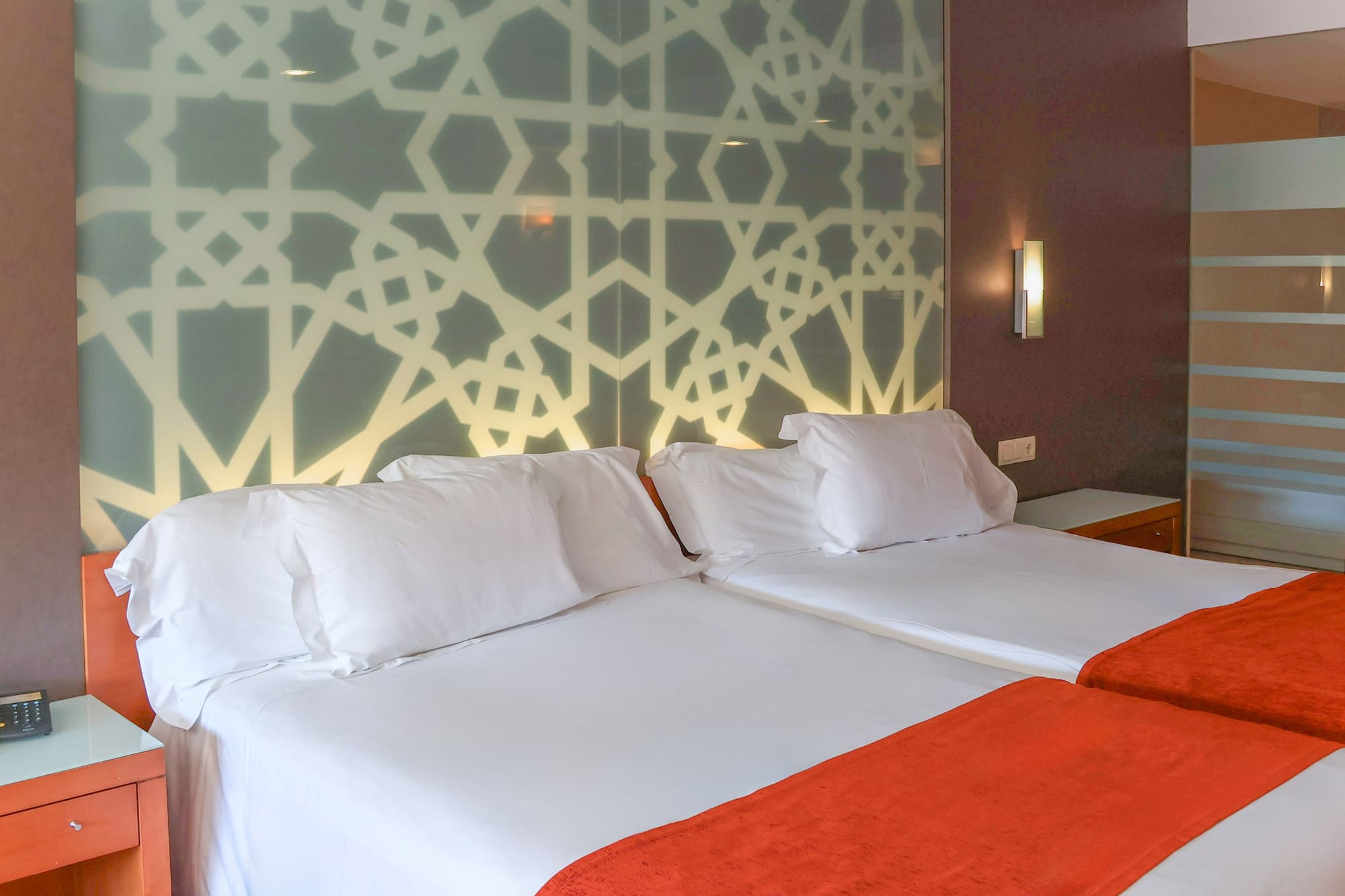 Hotel room with twin beds, white bedding, orange throws, and geometric headboard.