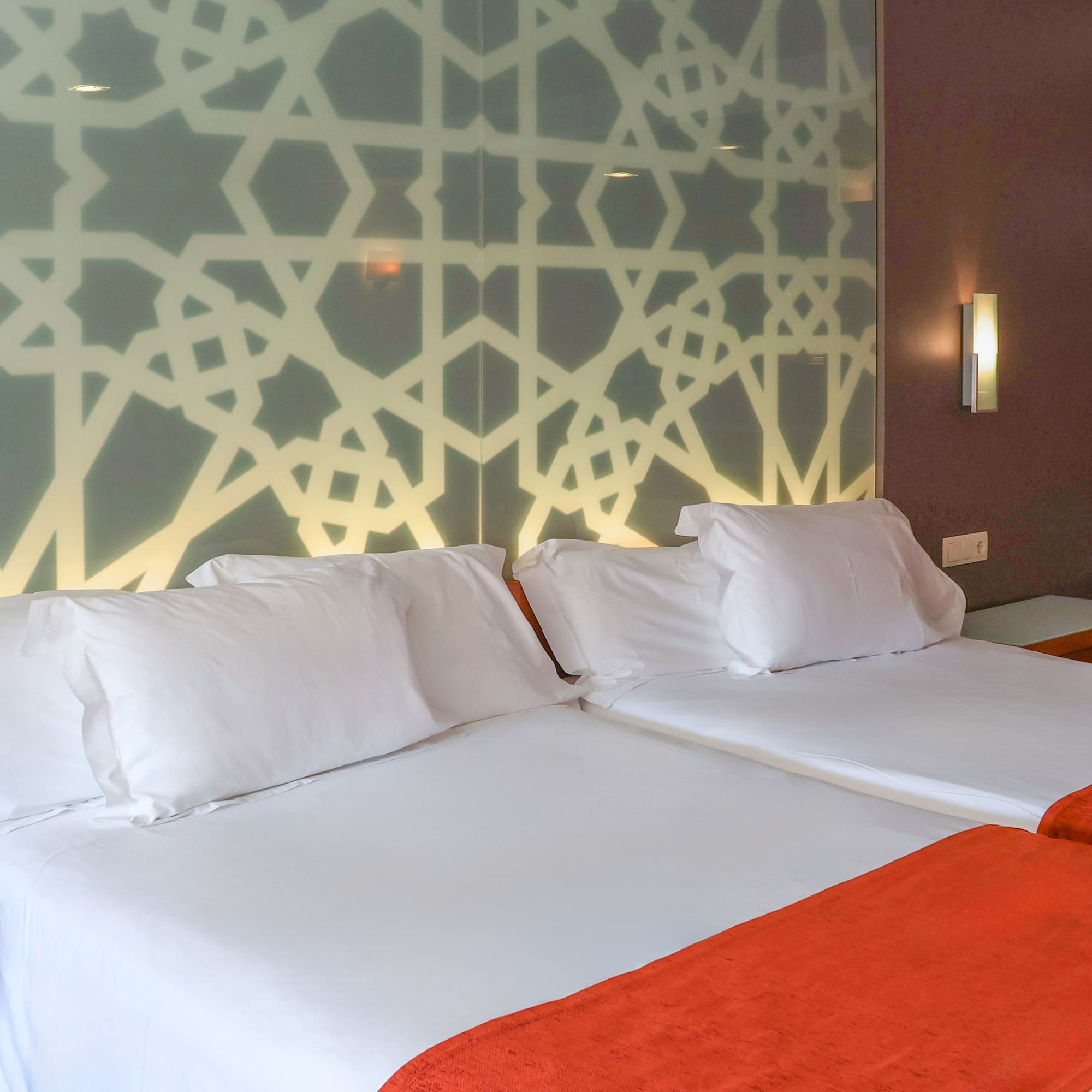 Hotel room with twin beds, white bedding, orange throws, and geometric headboard.