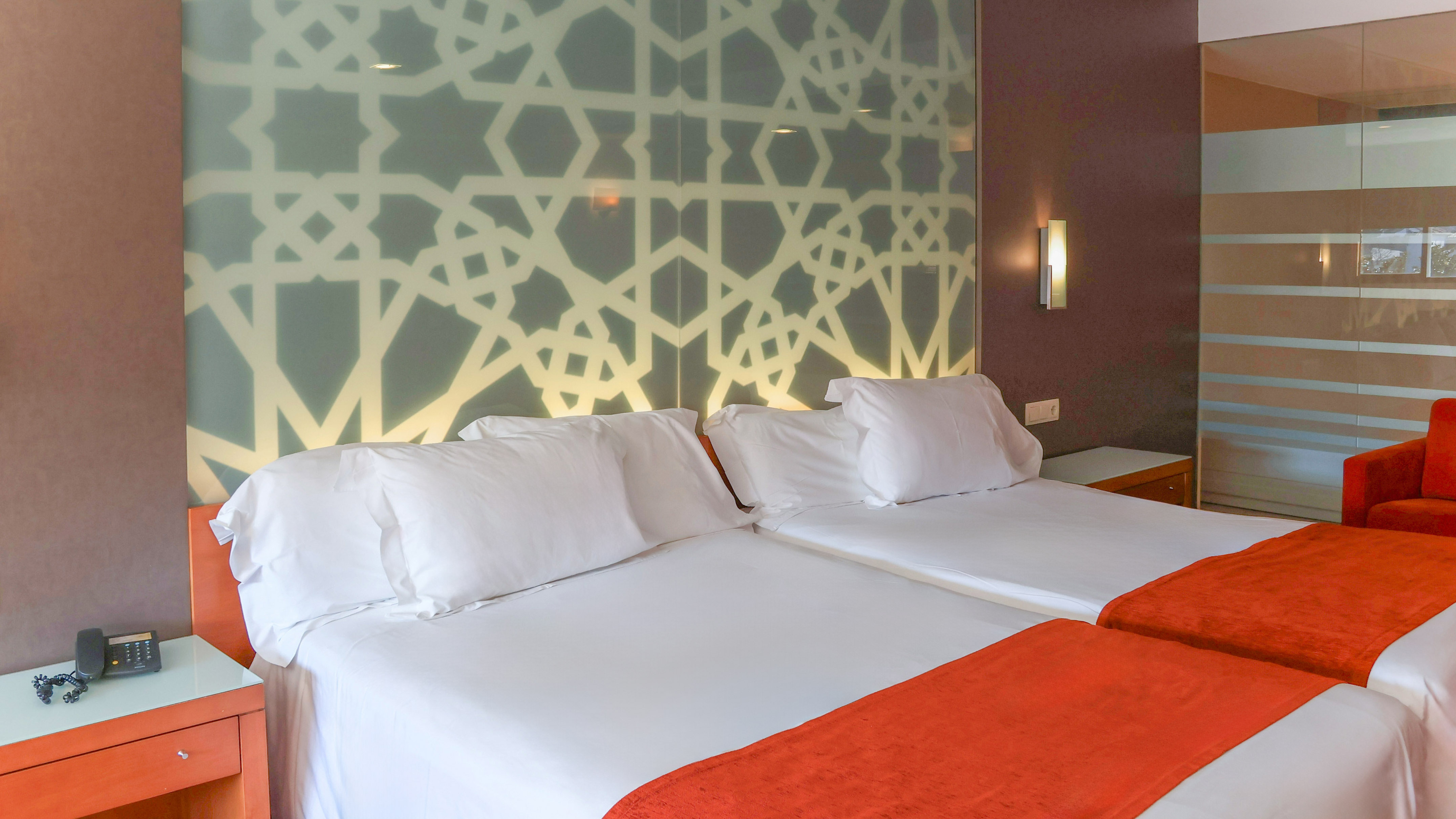 Hotel room with twin beds, white bedding, orange throws, and geometric headboard.