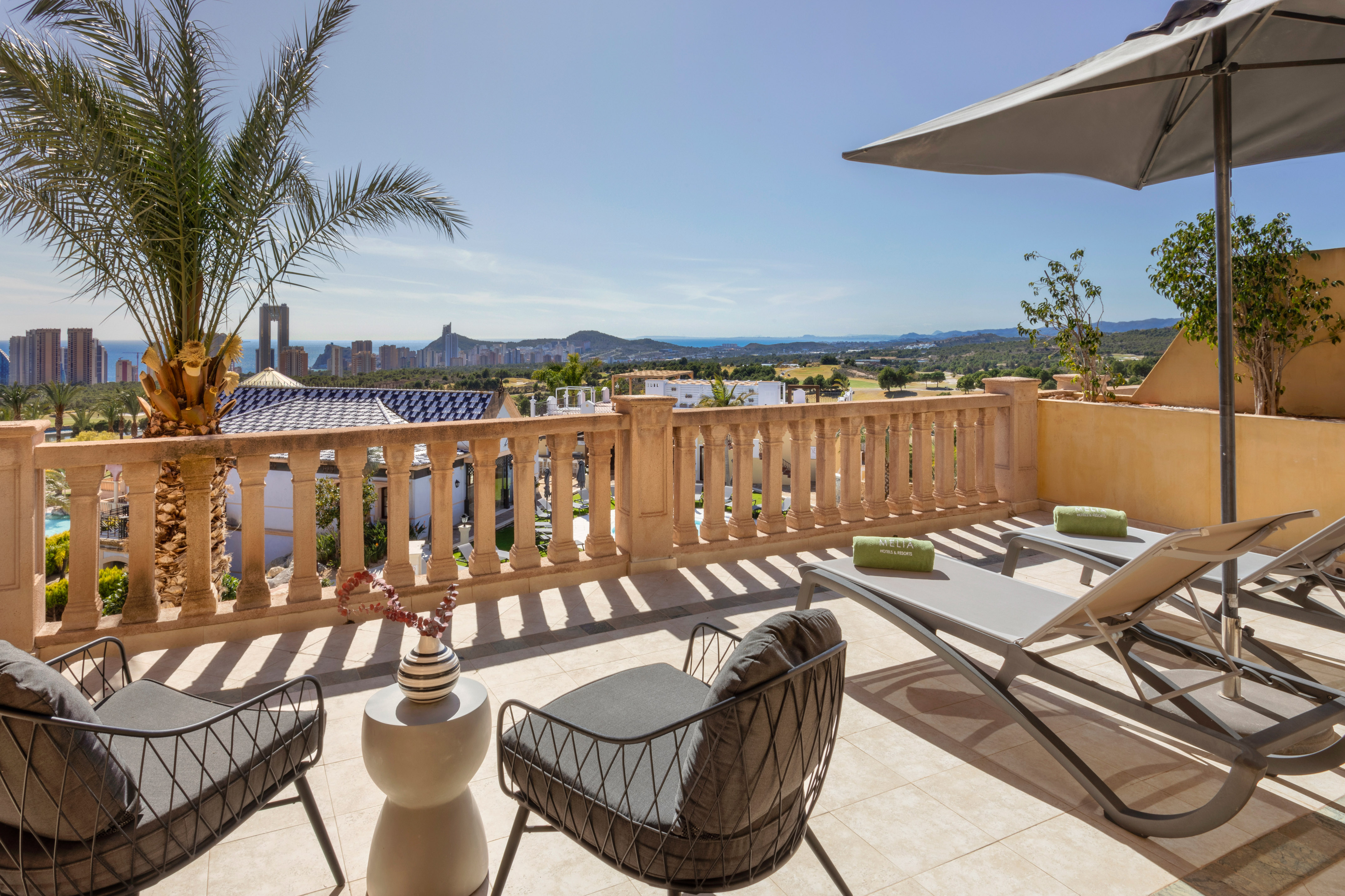 Resort terrace with ocean view, Benidorm skyline, palm tree, and sun loungers.