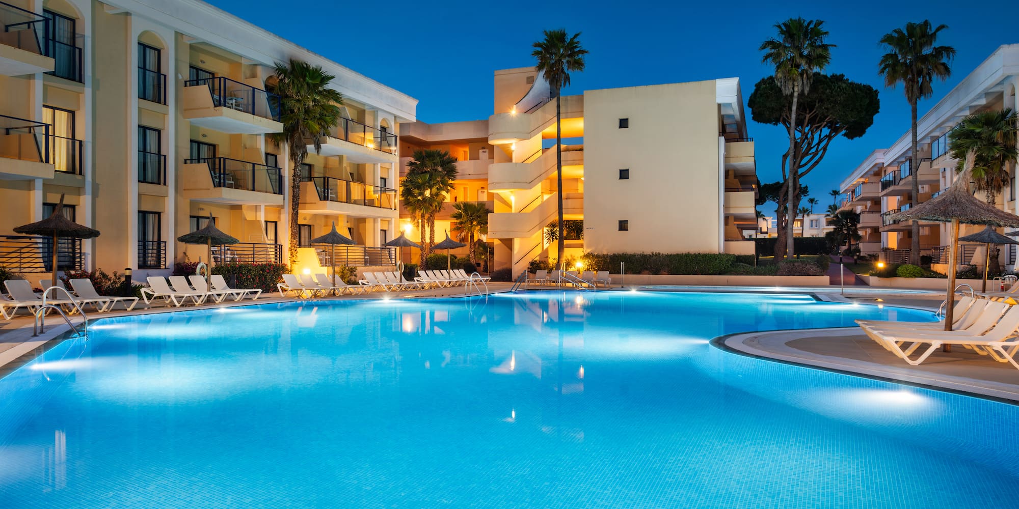 Illuminated hotel swimming pool at night with palm trees and lounge chairs.