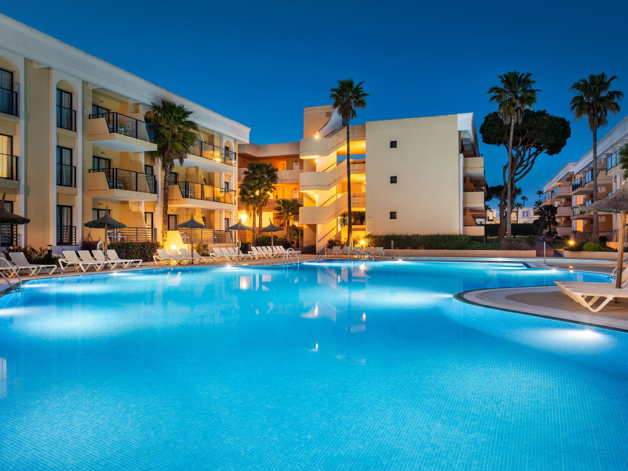 Illuminated resort pool at night, surrounded by hotel buildings and palm trees.