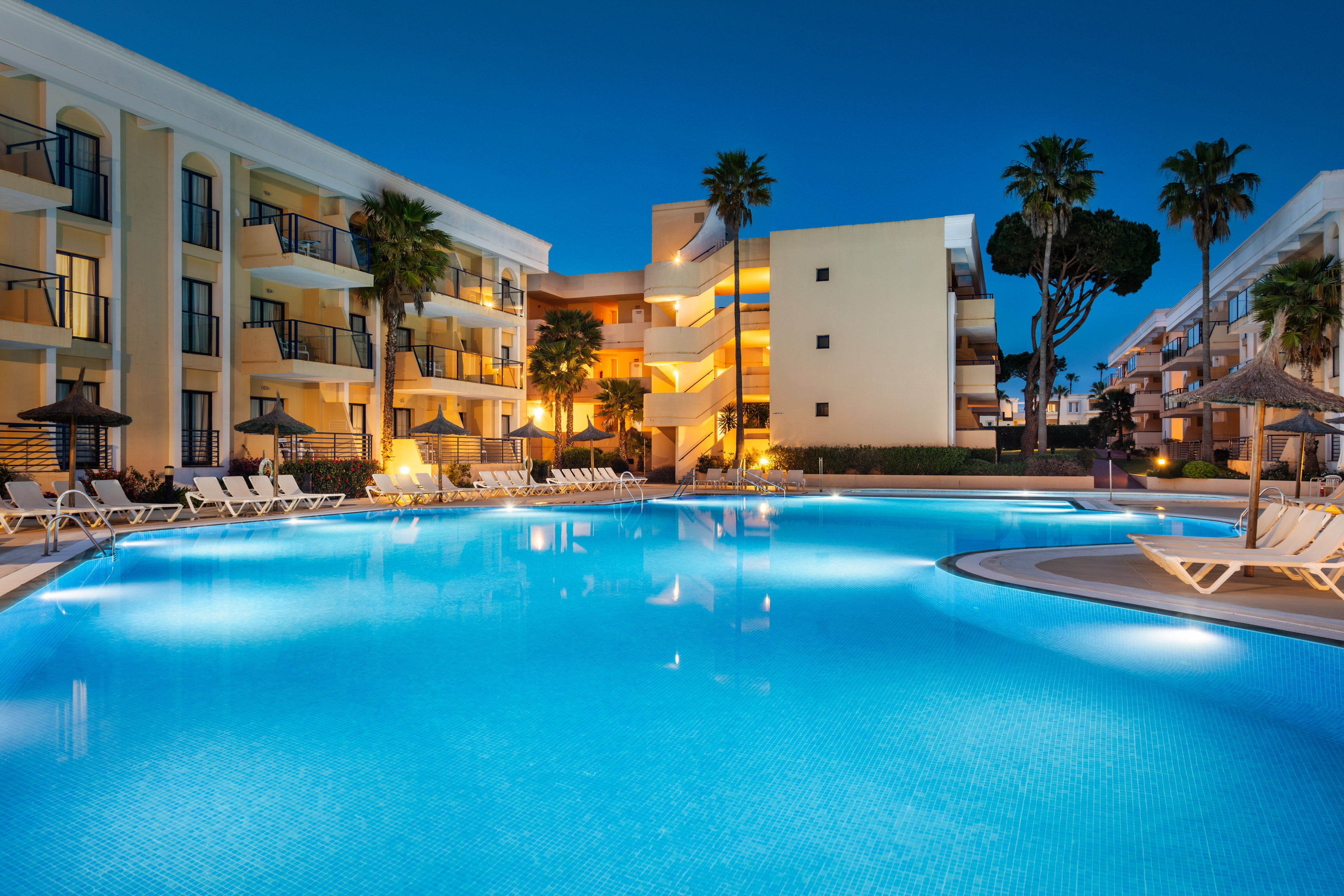 Illuminated hotel swimming pool at night with palm trees and lounge chairs.