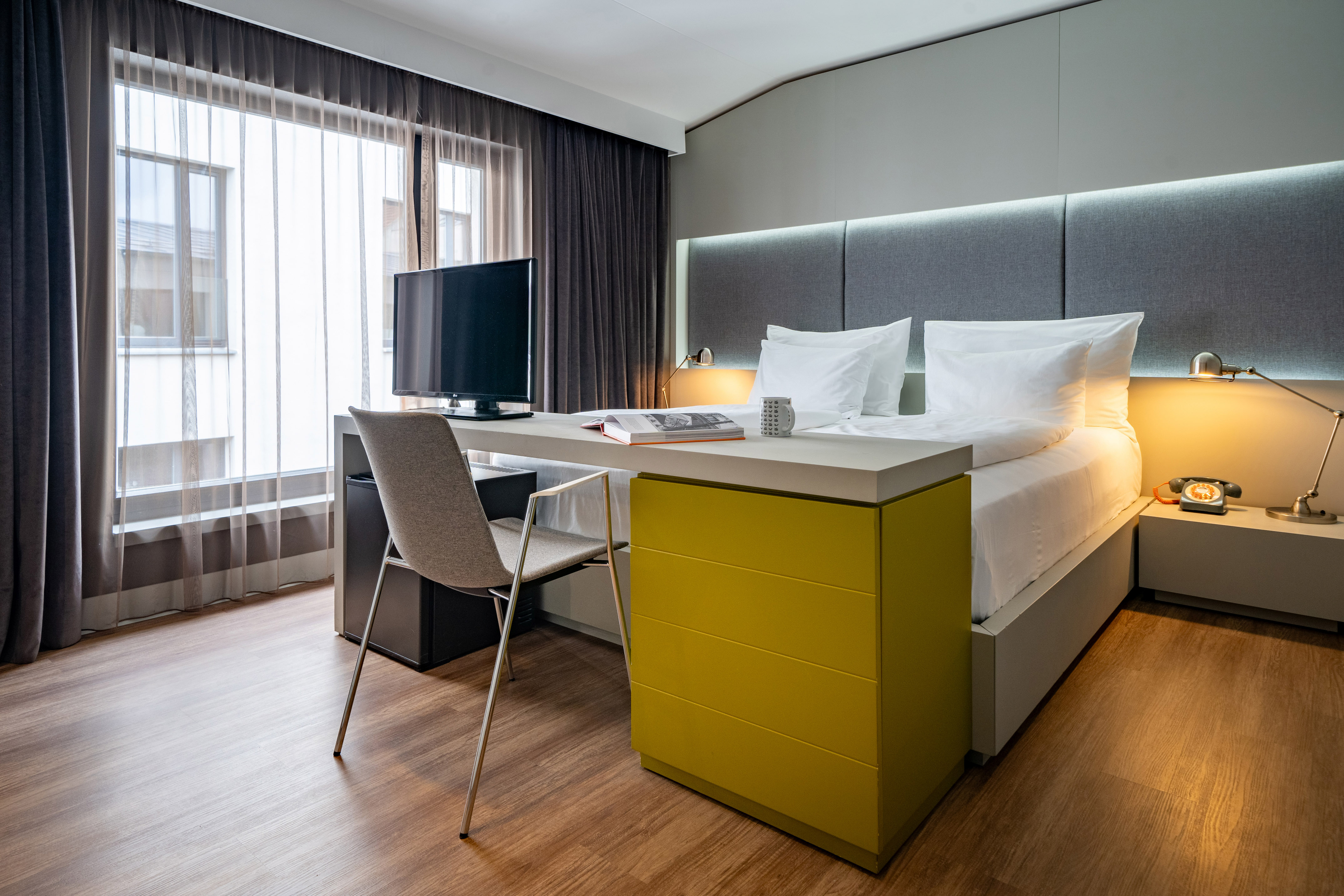 Modern hotel bedroom with bed, integrated desk, TV, and large window.