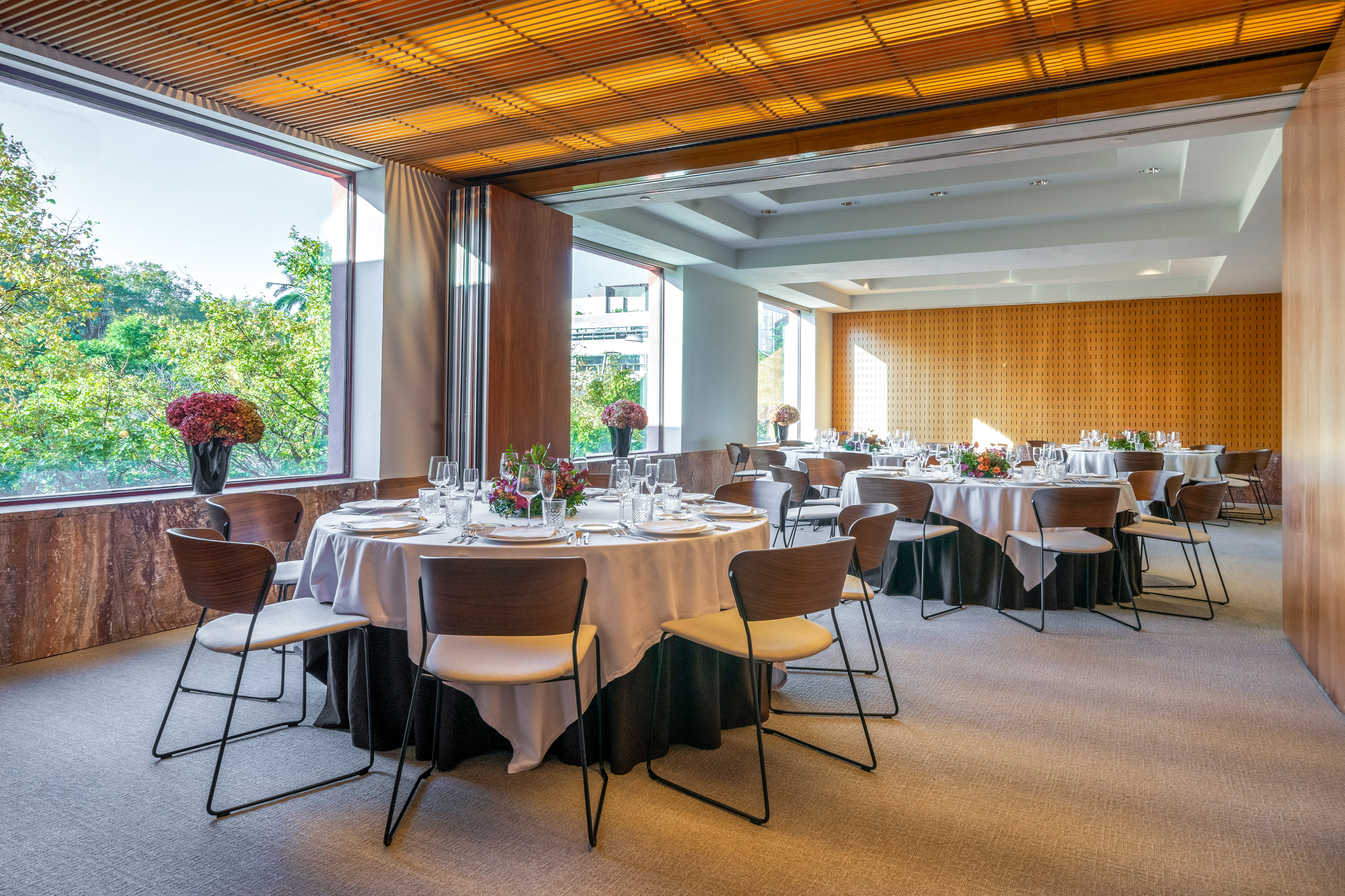 Modern event room with set dining tables, garden view, and wooden ceiling.