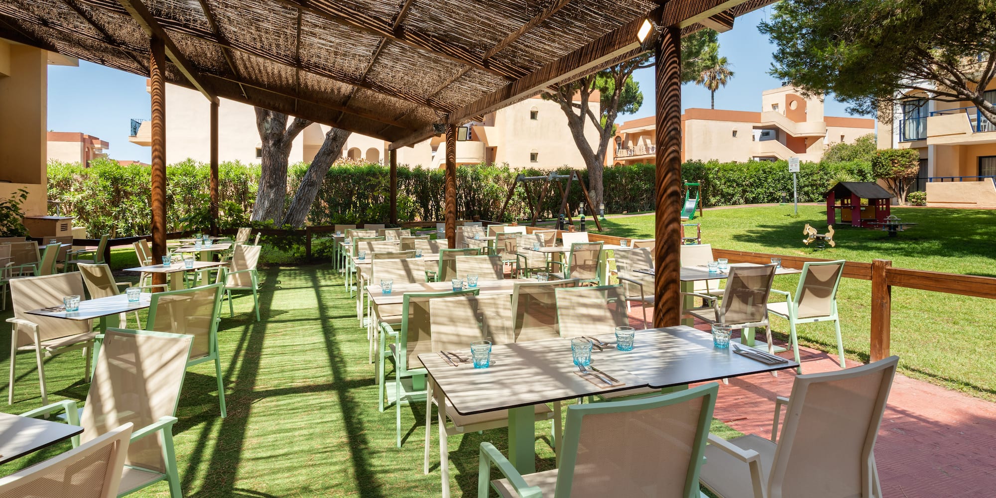 Shaded outdoor resort dining area, tables on green turf, overlooking garden.