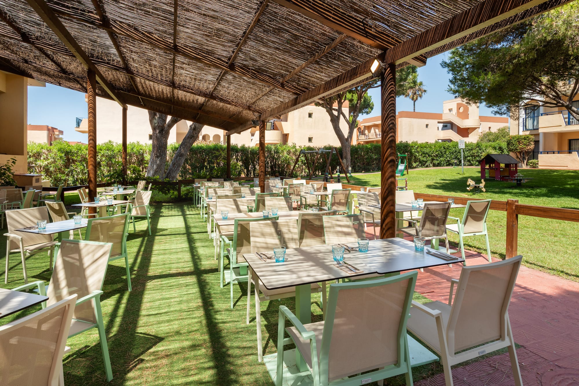 Shaded outdoor resort dining area, tables on green turf, overlooking garden.