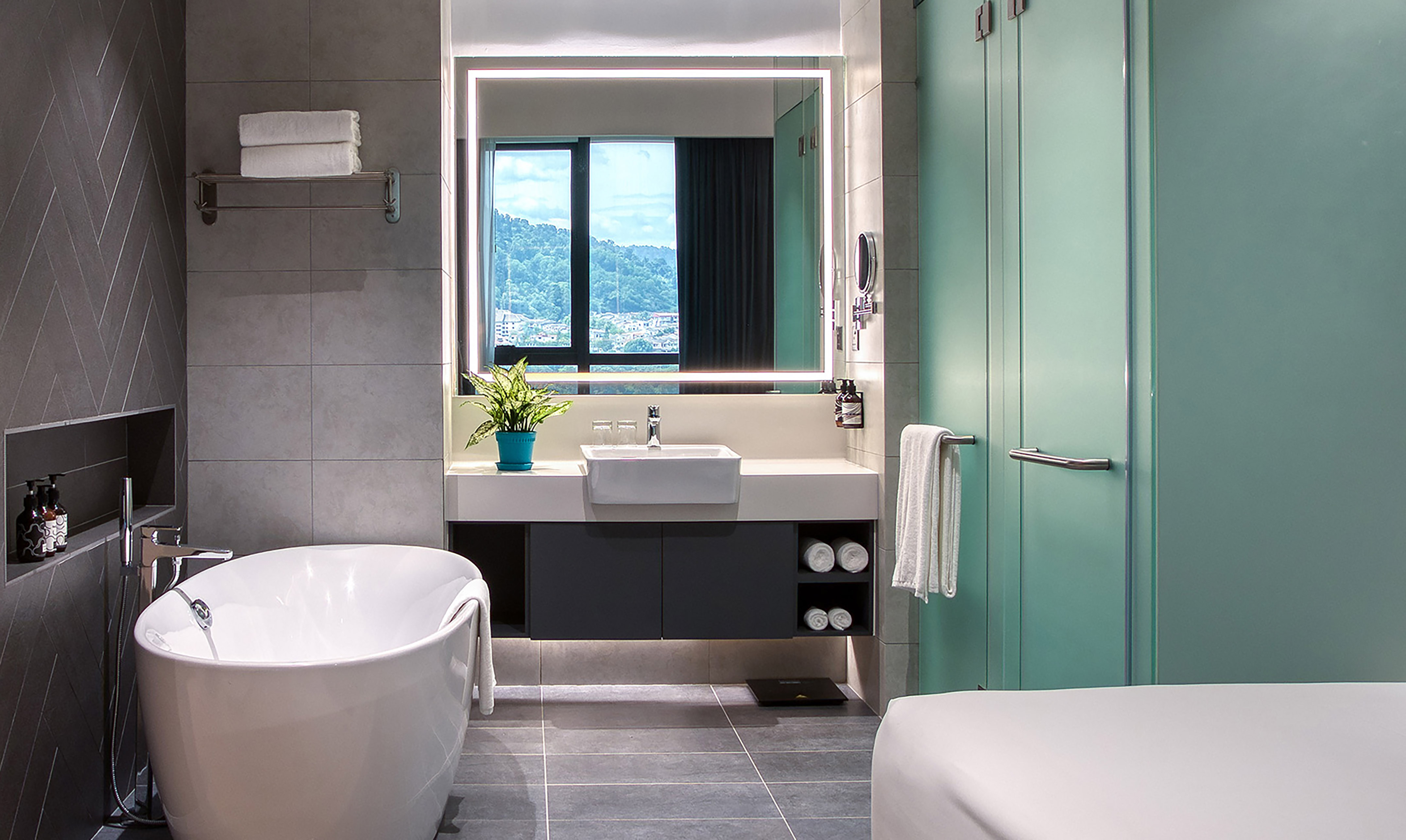 Hotel bathroom with freestanding tub, vanity, illuminated mirror, mountain view.