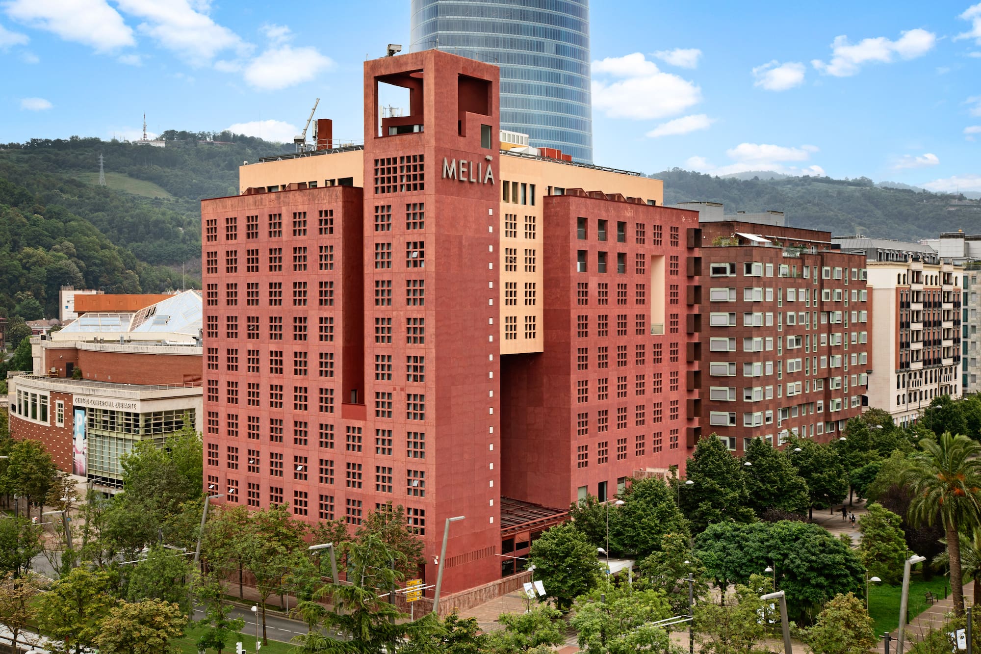 Modern red Melia hotel building with grid windows, glass skyscraper, and urban trees.