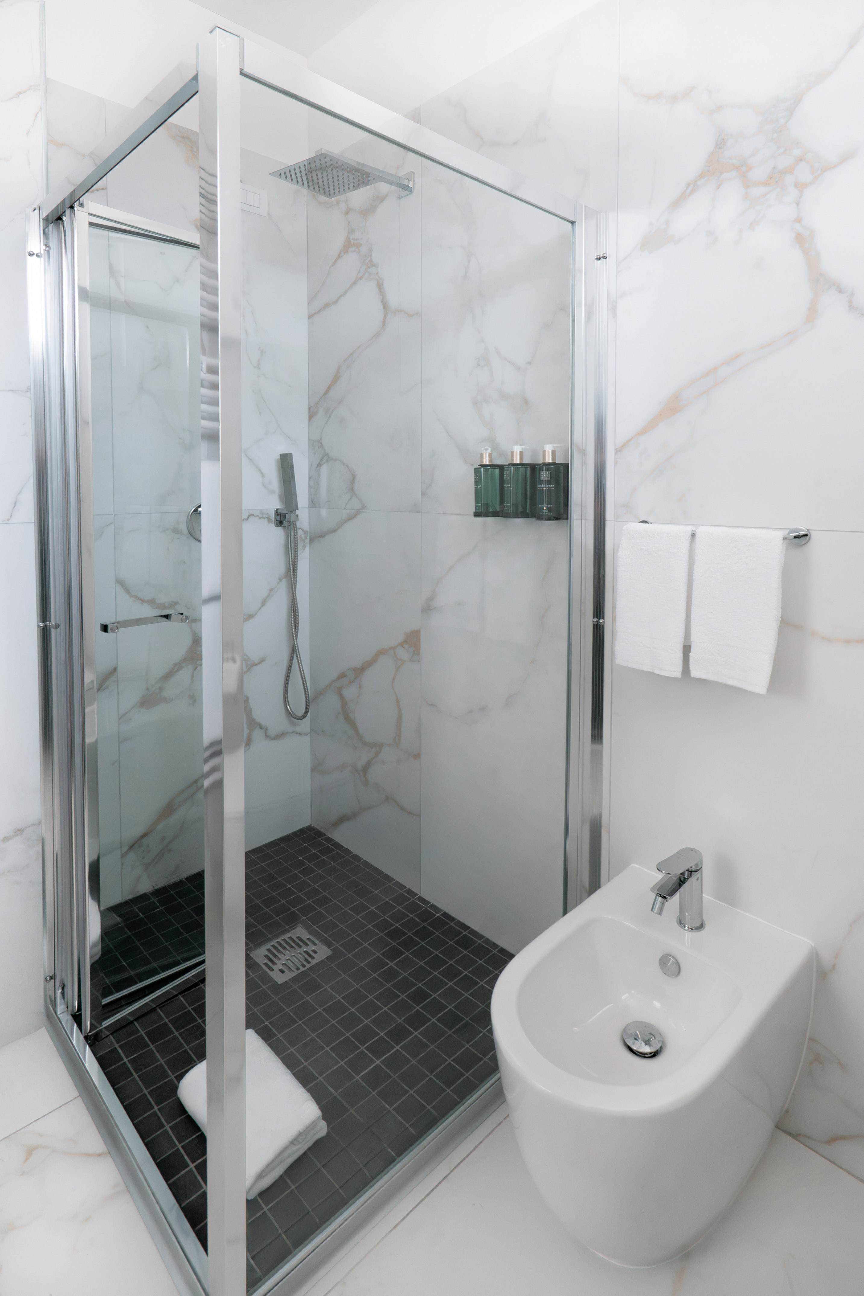 Modern bathroom with glass shower, white marble walls, dark floor, and bidet.