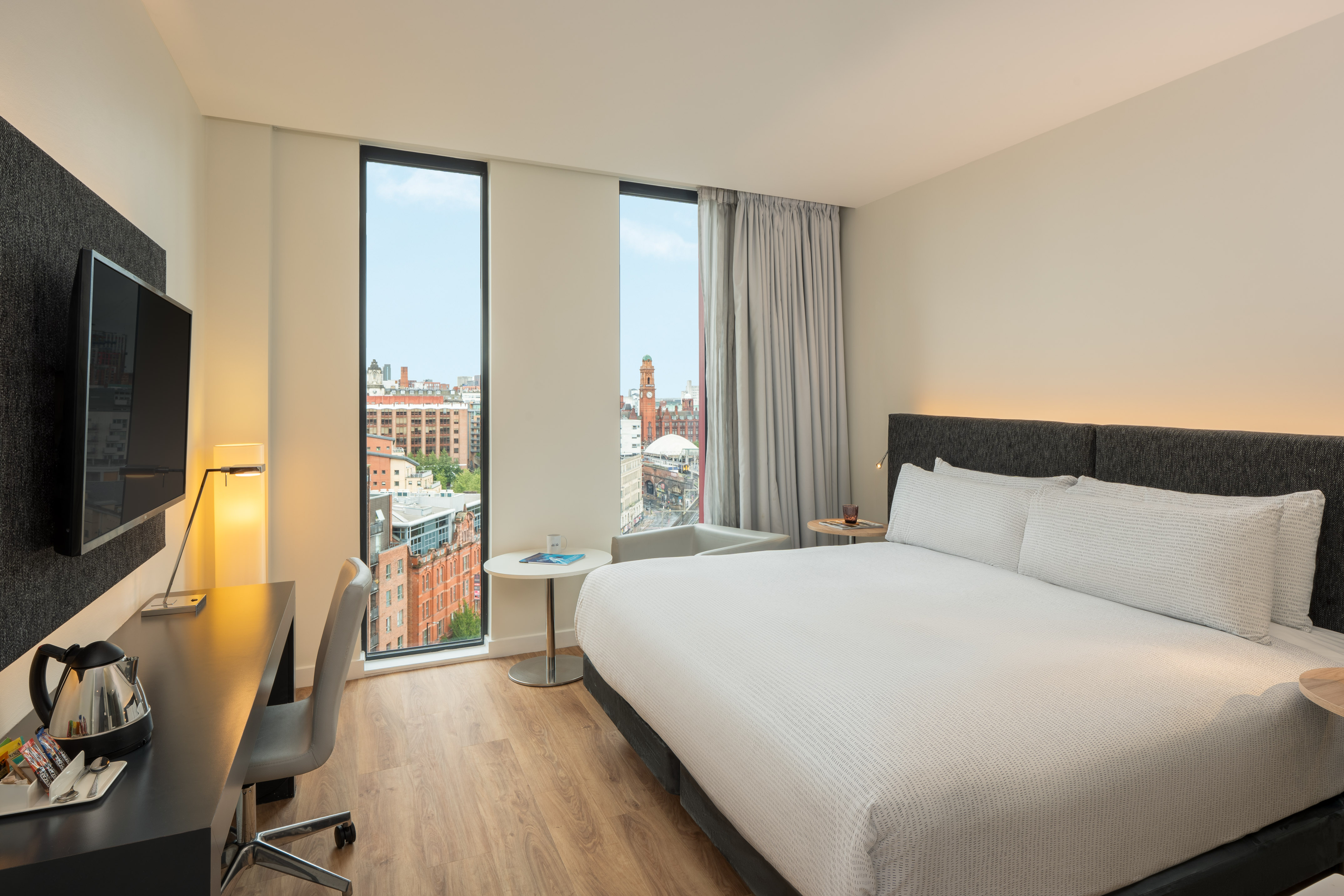 Modern hotel room with large bed, desk, TV, and city view through tall windows.