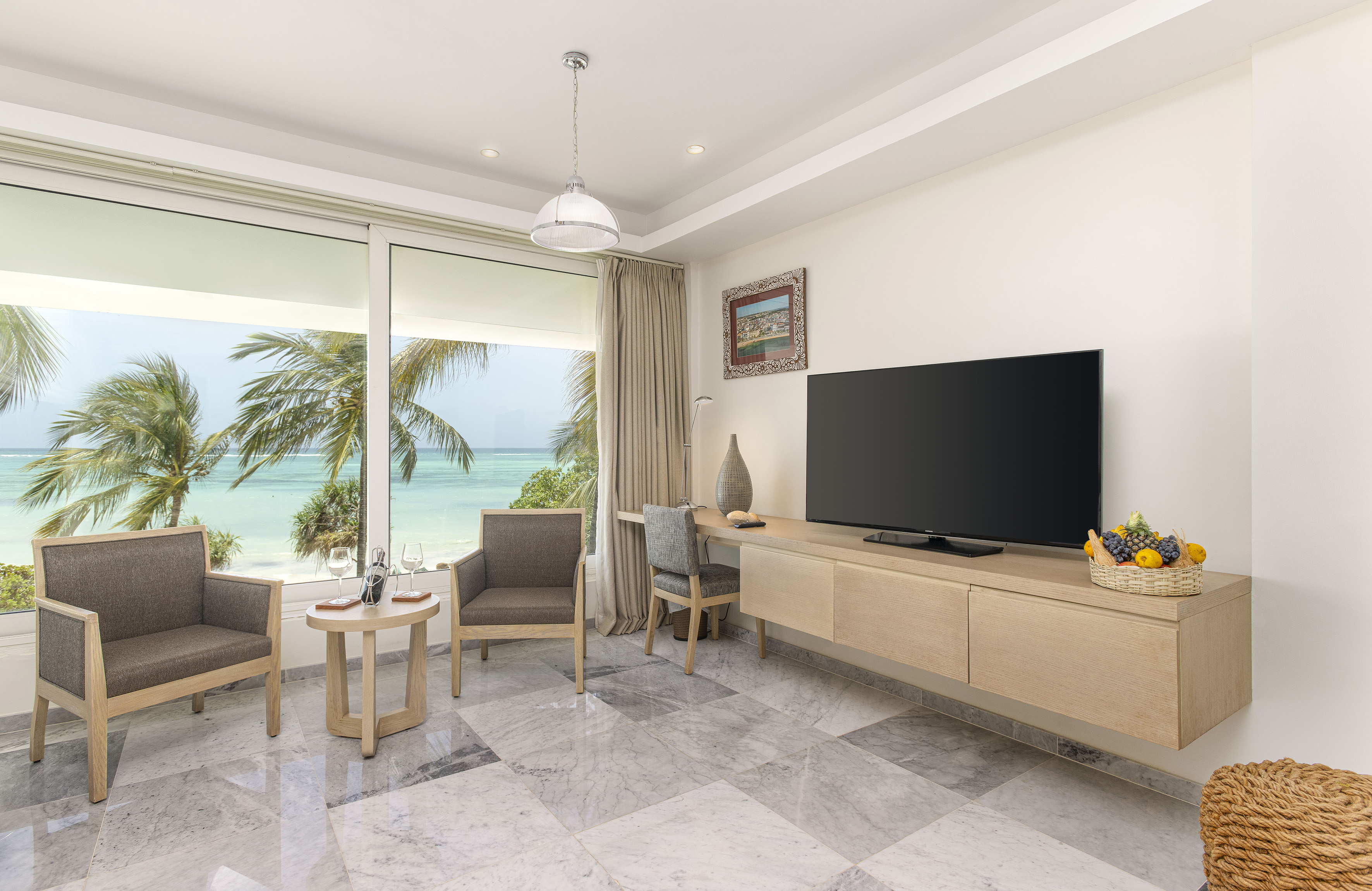 Tropical resort living room with ocean view, palm trees, TV, and armchairs.