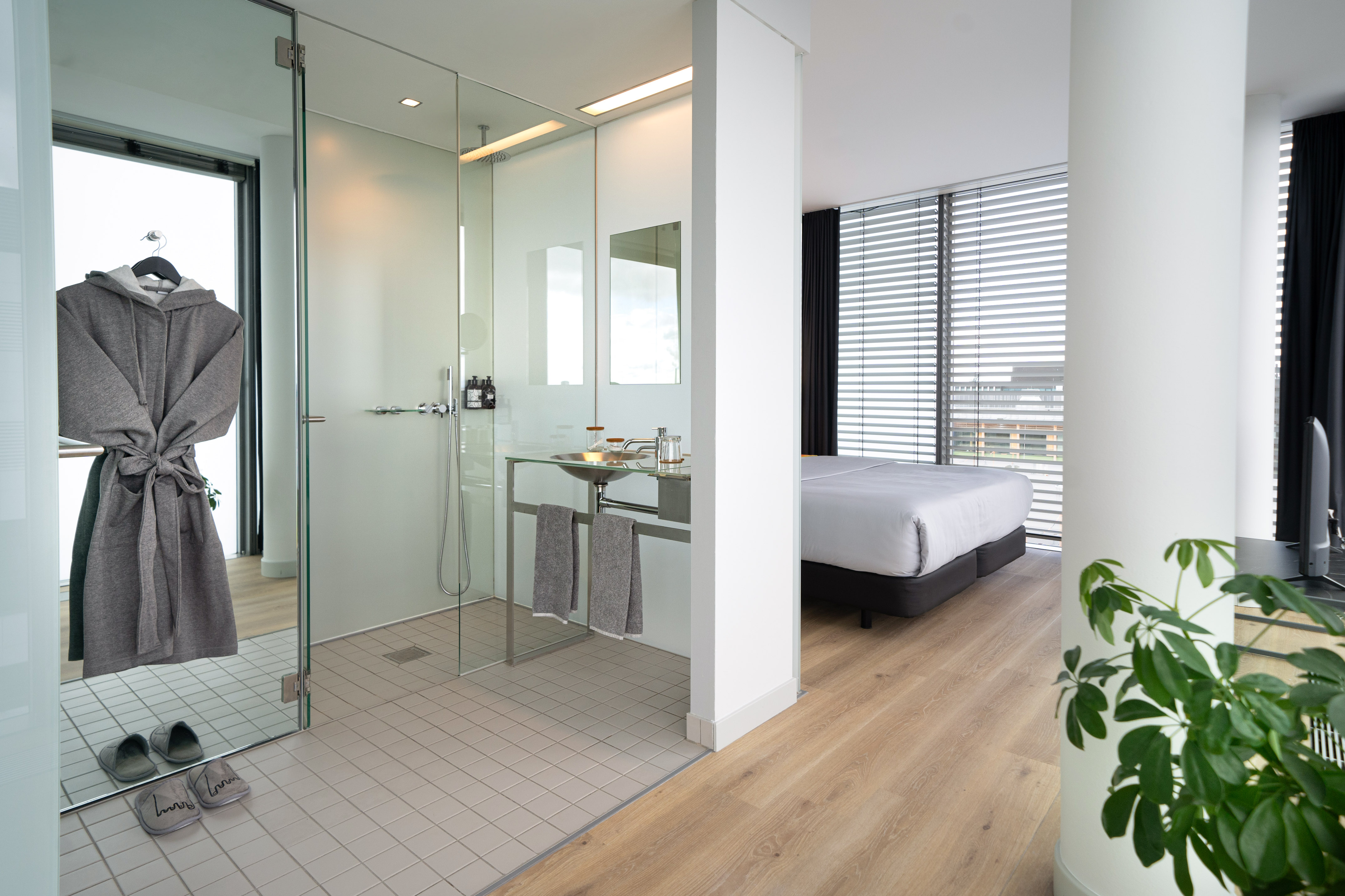 Modern hotel room with open bathroom, glass shower, vanity, and bed.
