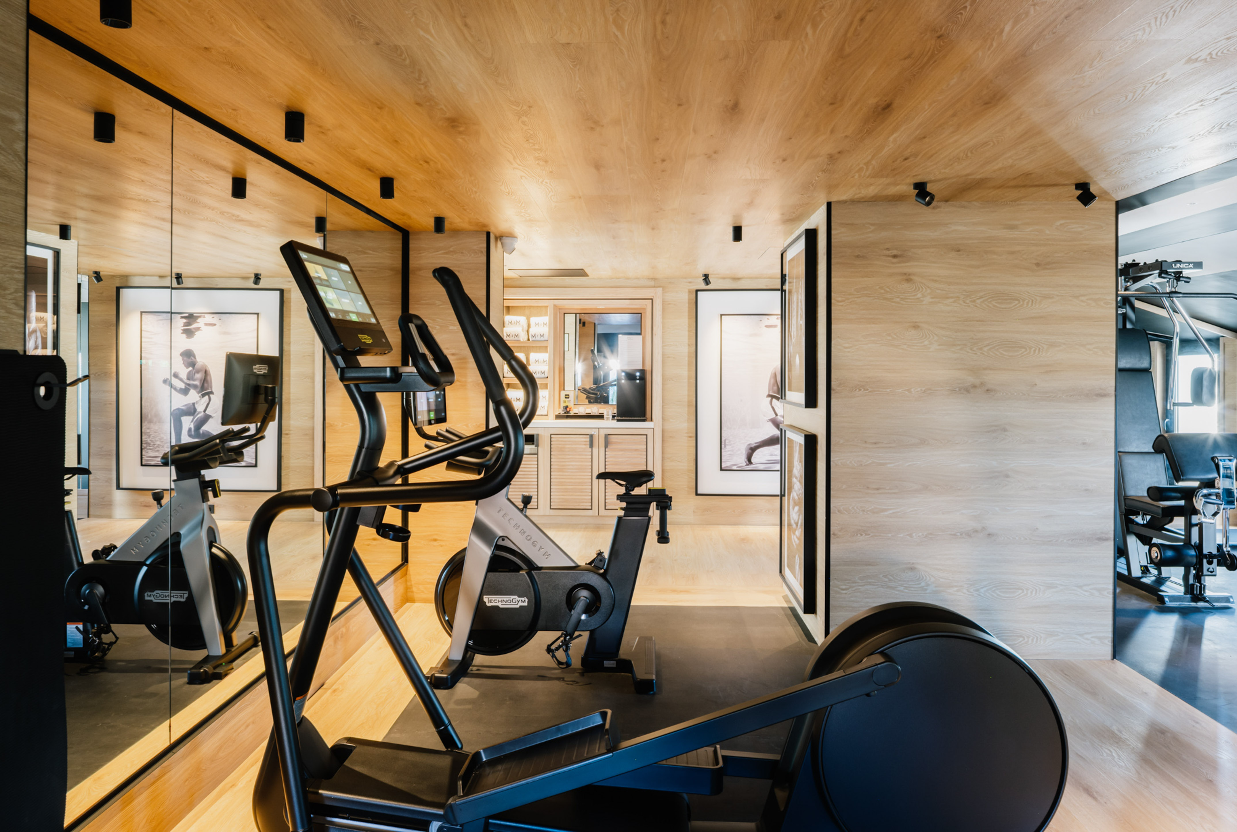 Modern fitness room with Technogym equipment, wood accents, and mirrored wall.