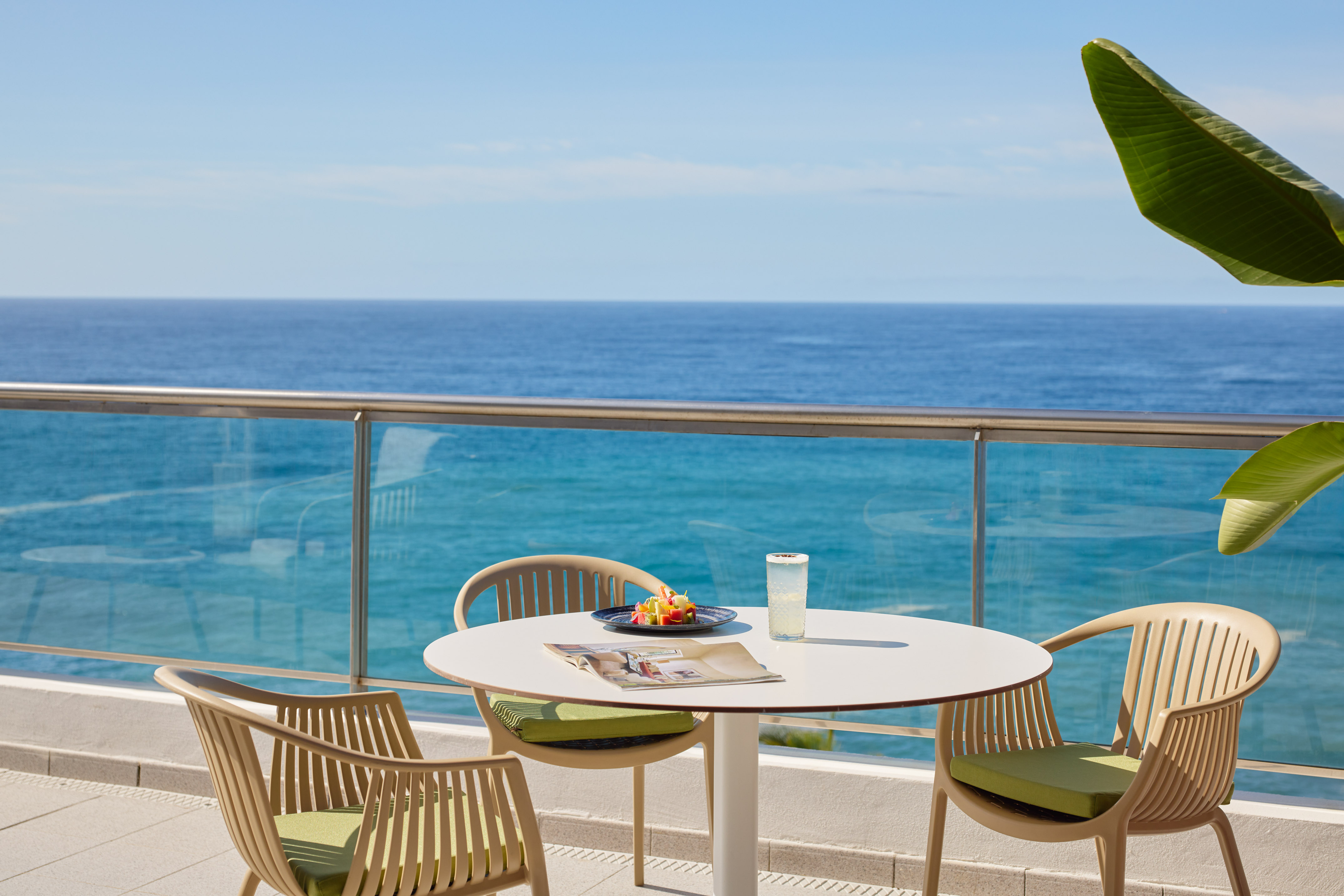 Sunny balcony with ocean view, table, chairs, fruit, and refreshing drink.