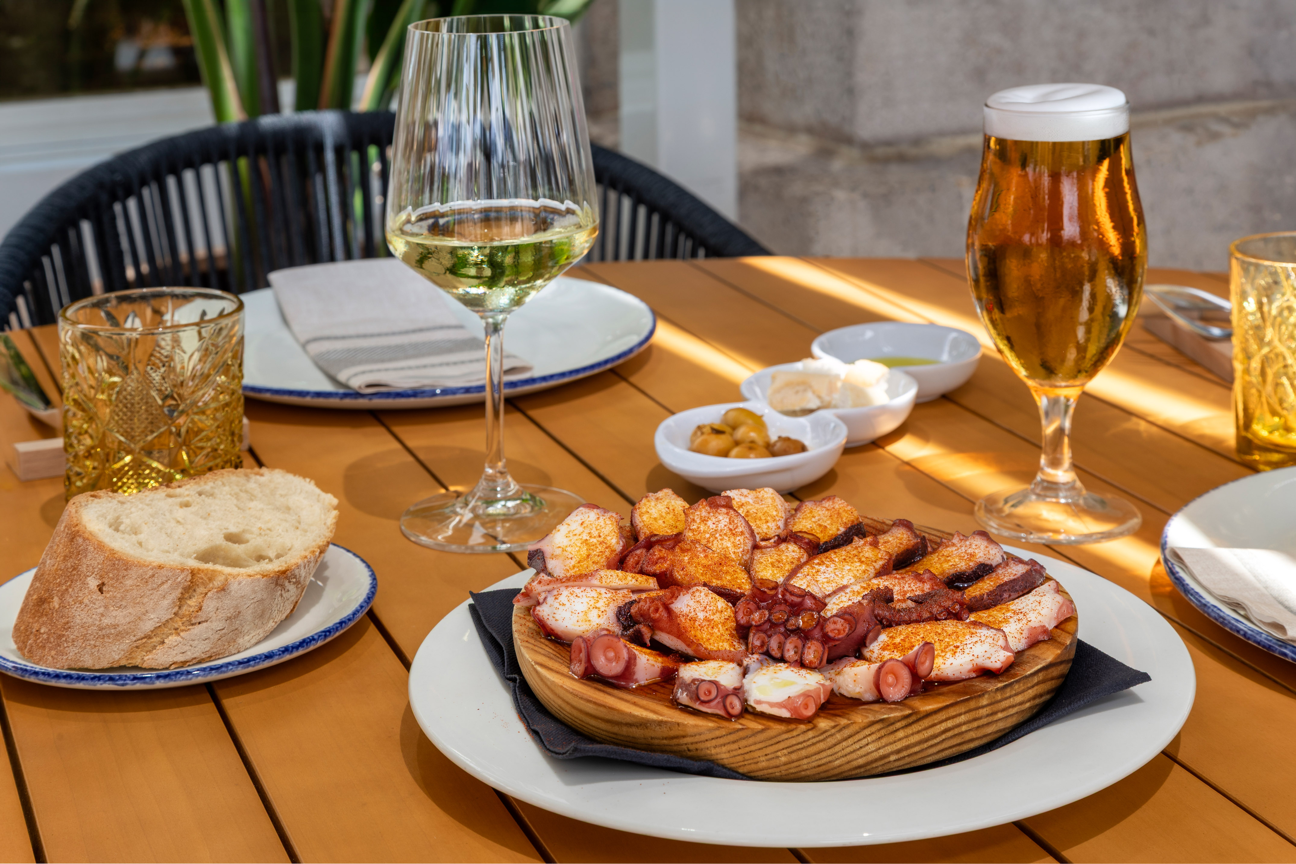 Pulpo a la gallega, pan, vino blanco y cerveza en mesa de madera exterior.