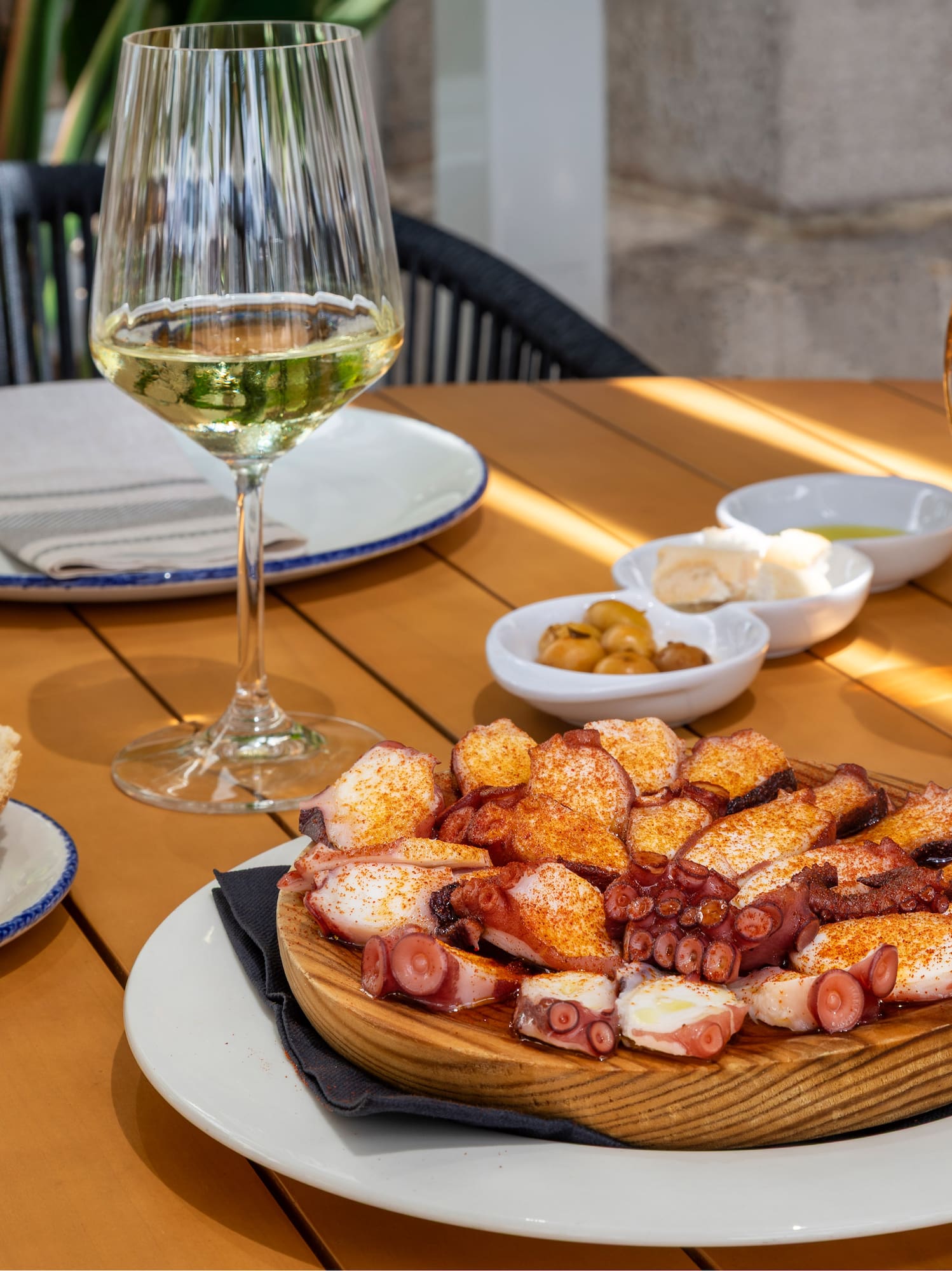 Pulpo a la gallega, pan, vino blanco y cerveza en mesa de madera exterior.