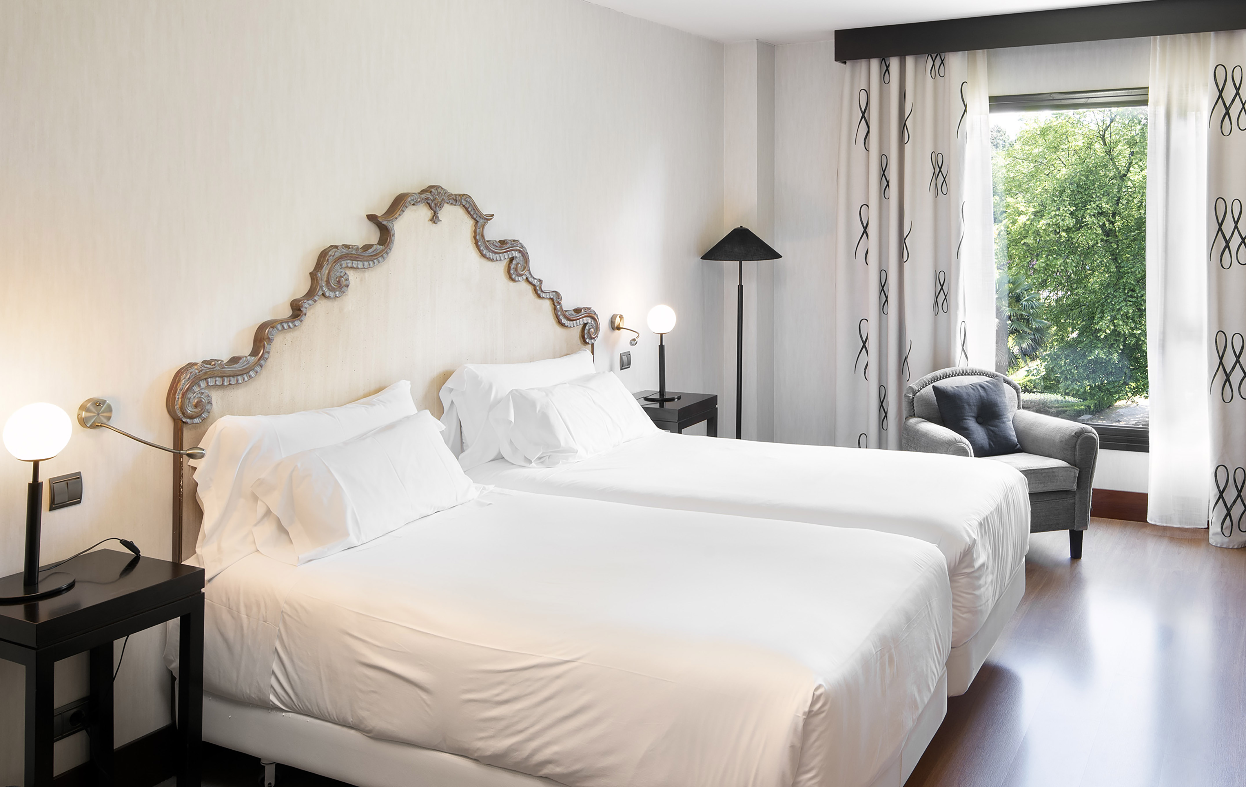 Hotel room with twin beds, carved headboard, and green outdoor view.