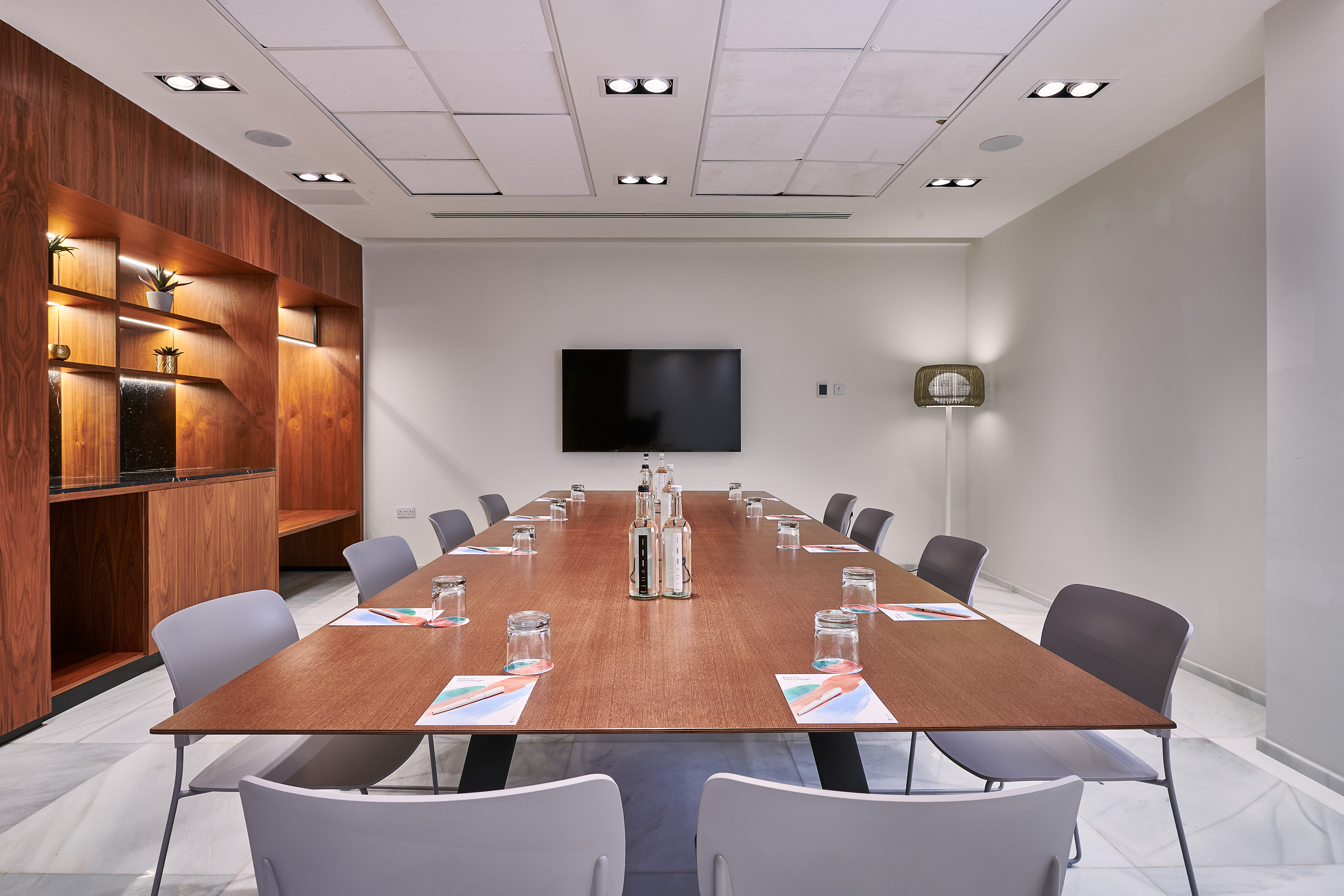 Modern meeting room with wooden table, grey chairs, presentation TV, and lit shelving.