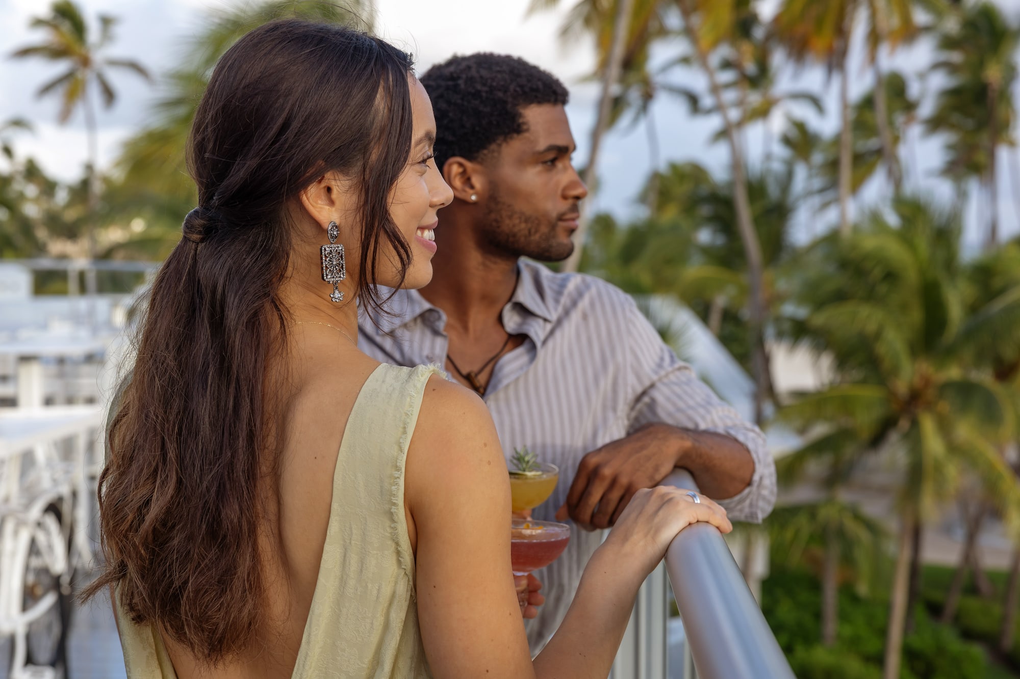 Couple with cocktails relaxing on a tropical balcony overlooking palm trees.
