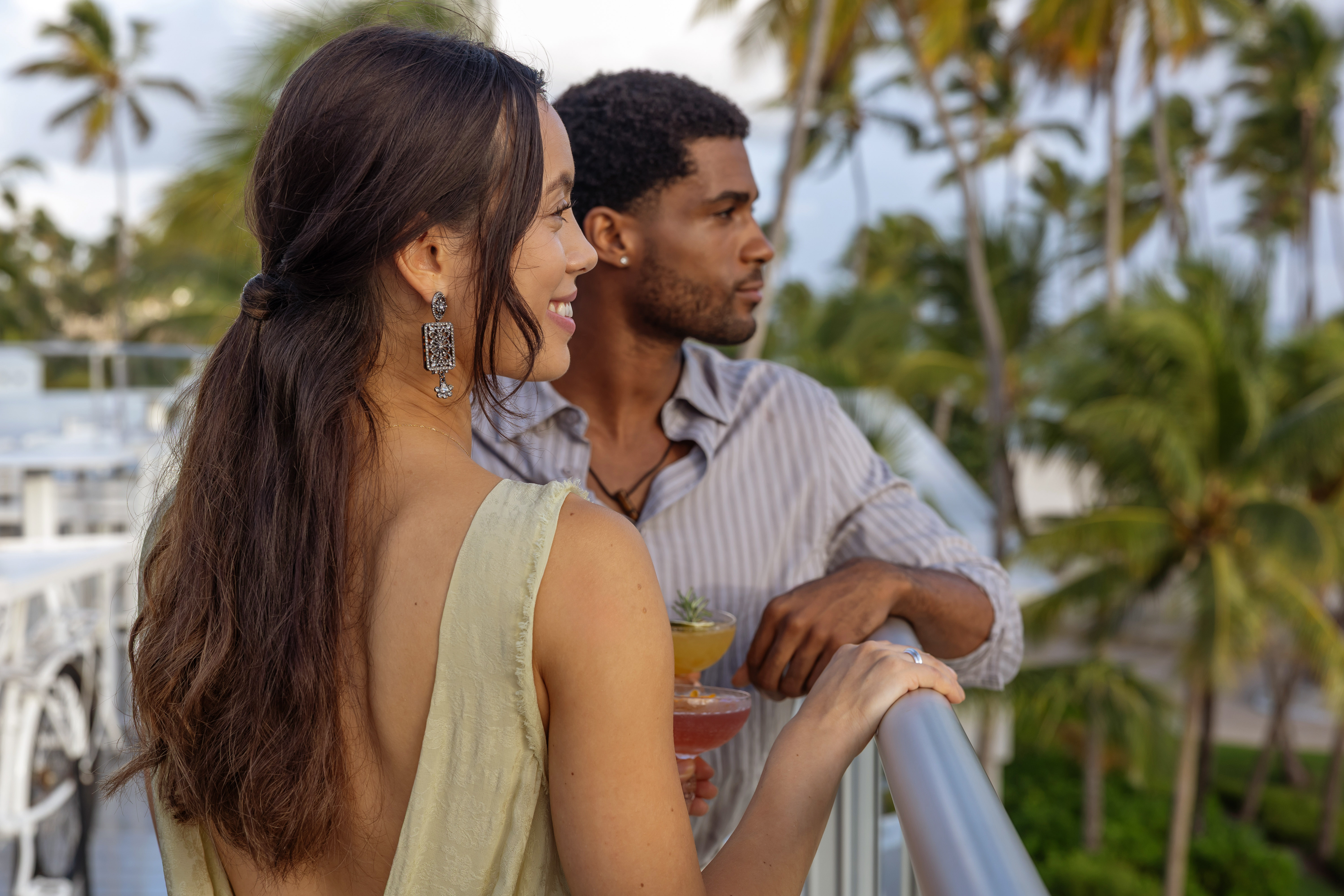 Couple with cocktails relaxing on a tropical balcony overlooking palm trees.