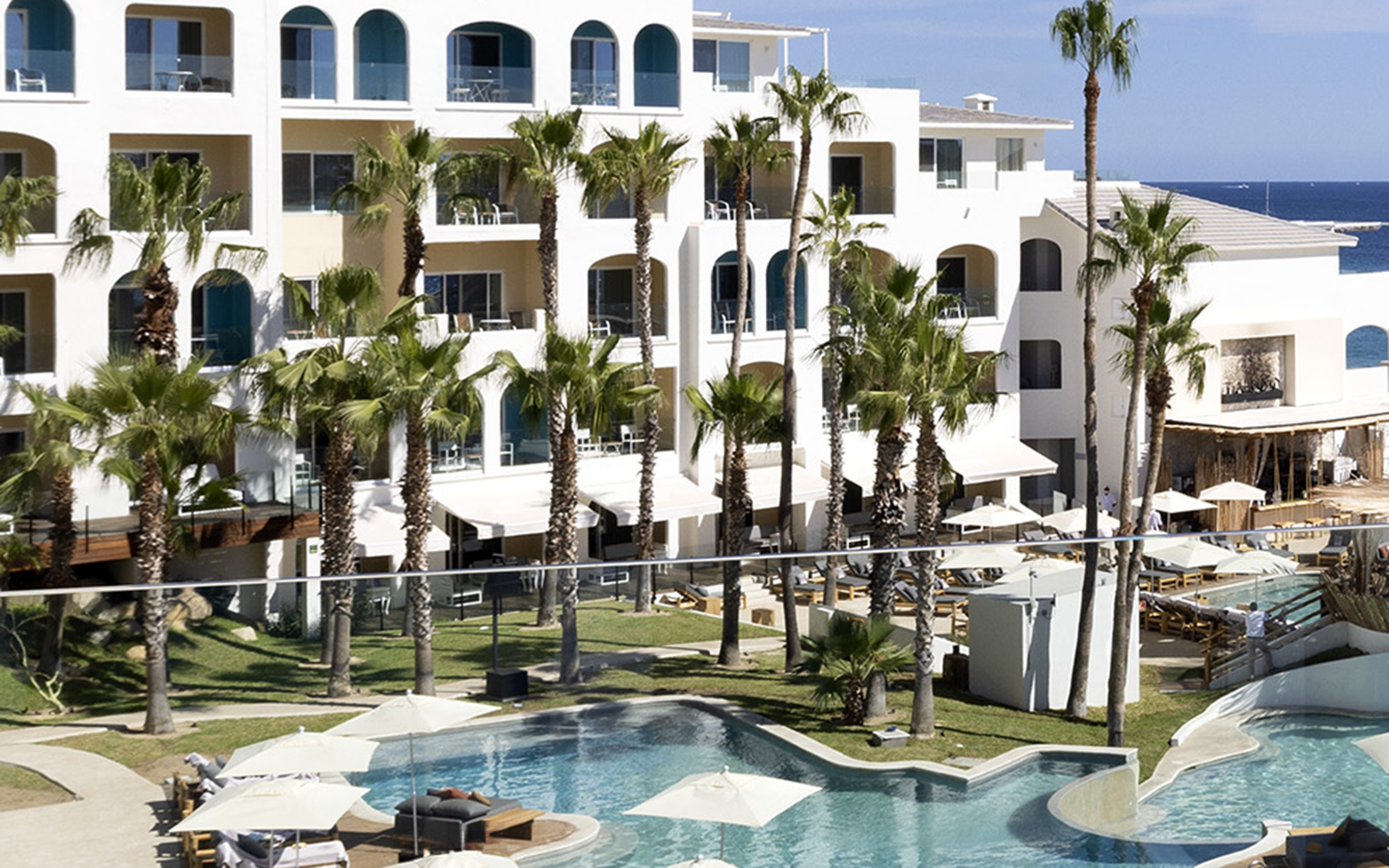 Tropical resort with white buildings, lush palm trees, swimming pools, and ocean.