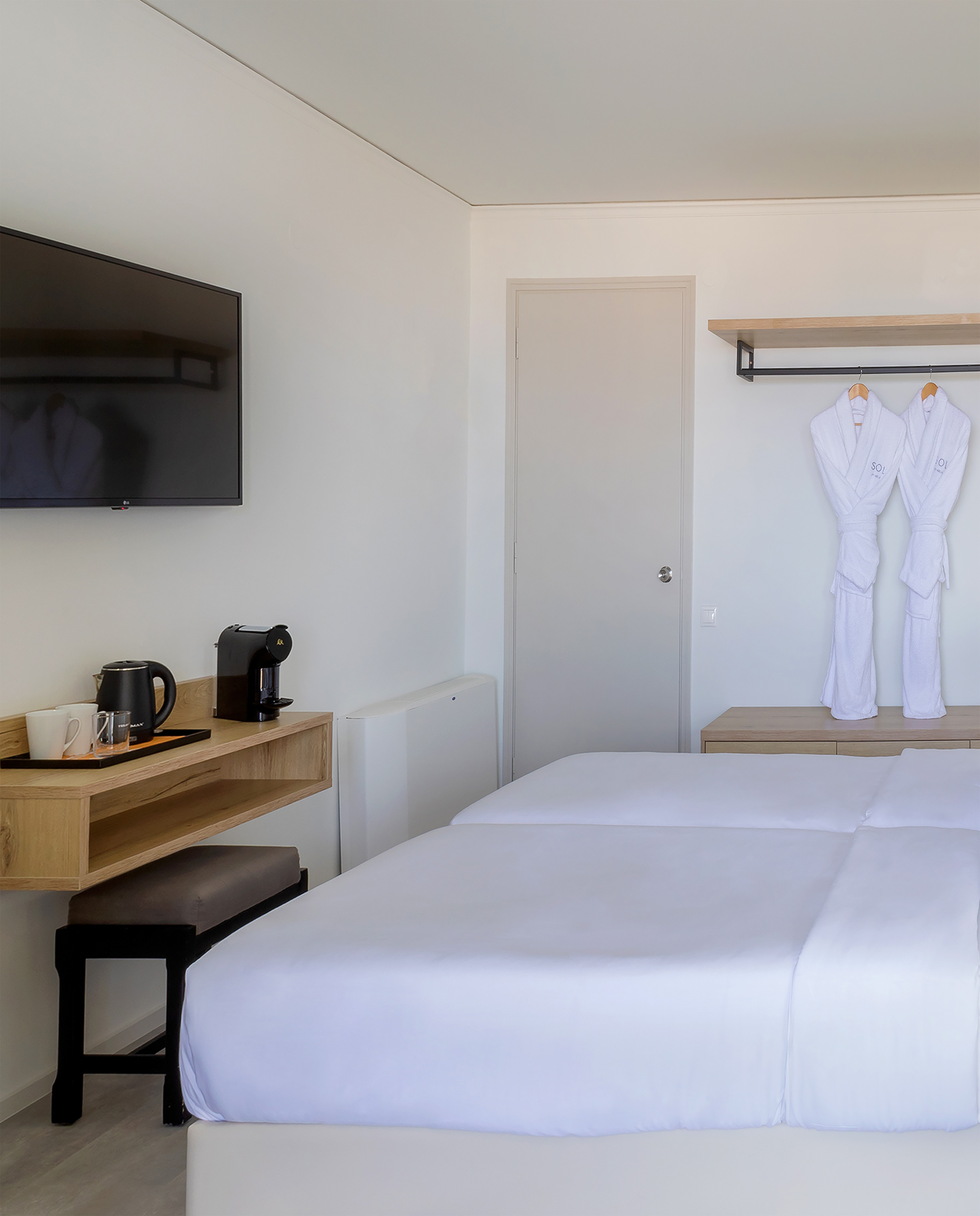 Modern hotel room with twin beds, TV, coffee maker, and bathrobes on wall rack.