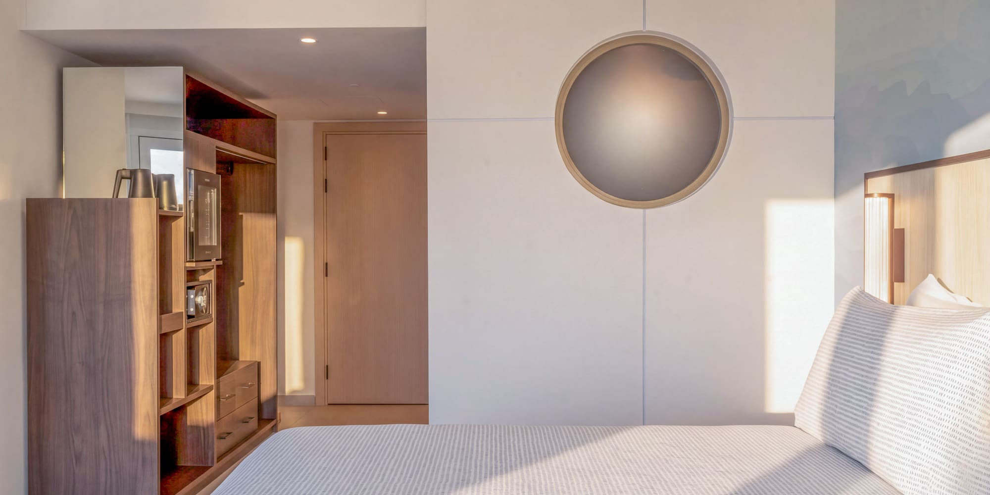 Modern hotel bedroom with bed, wooden storage, and circular wall light.