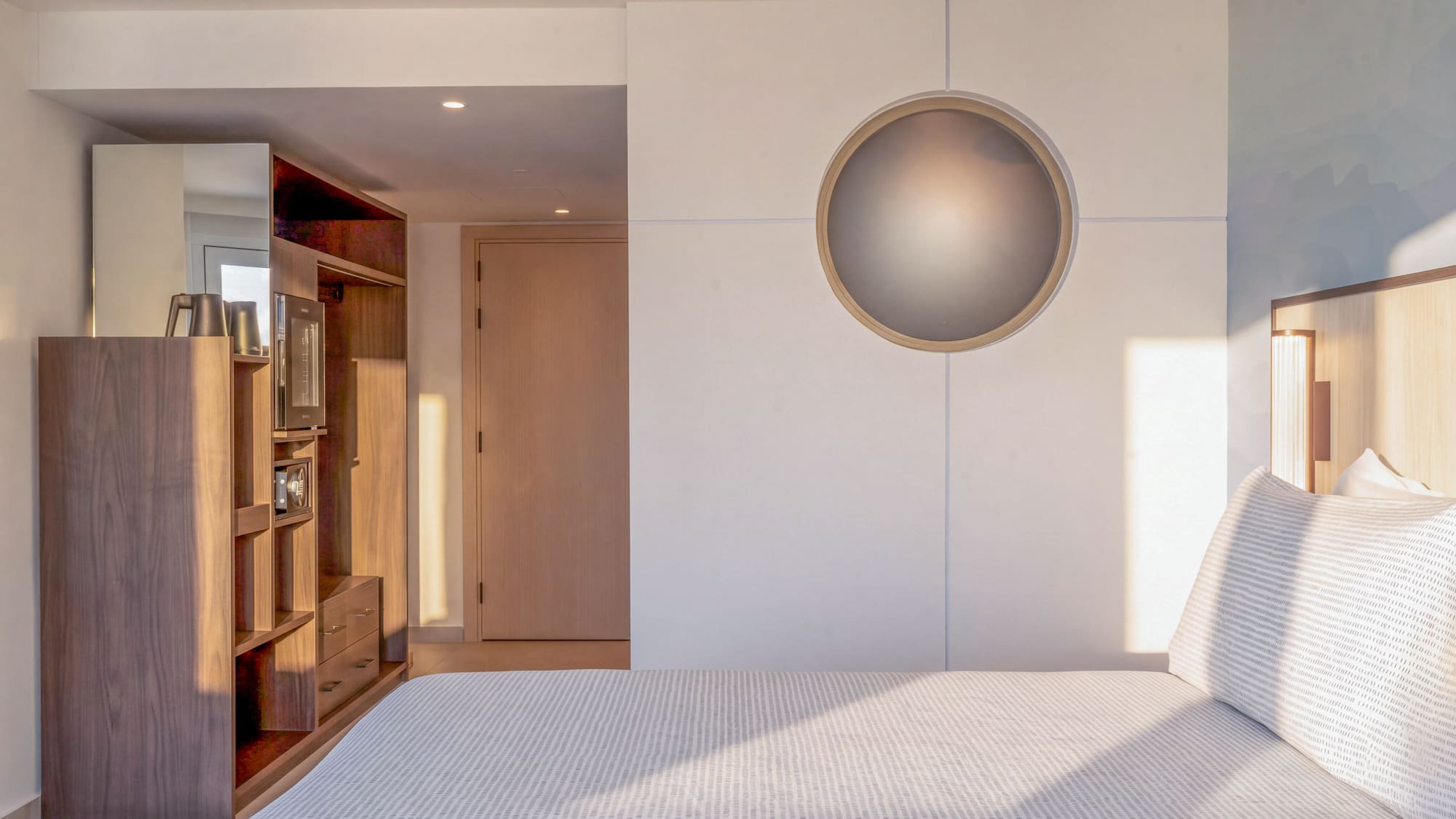 Modern hotel bedroom with bed, wooden storage, and circular wall light.