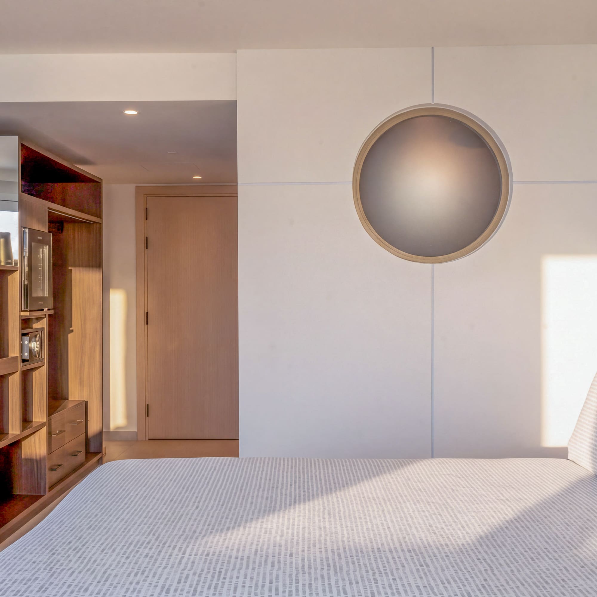 Modern hotel bedroom with bed, wooden storage, and circular wall light.