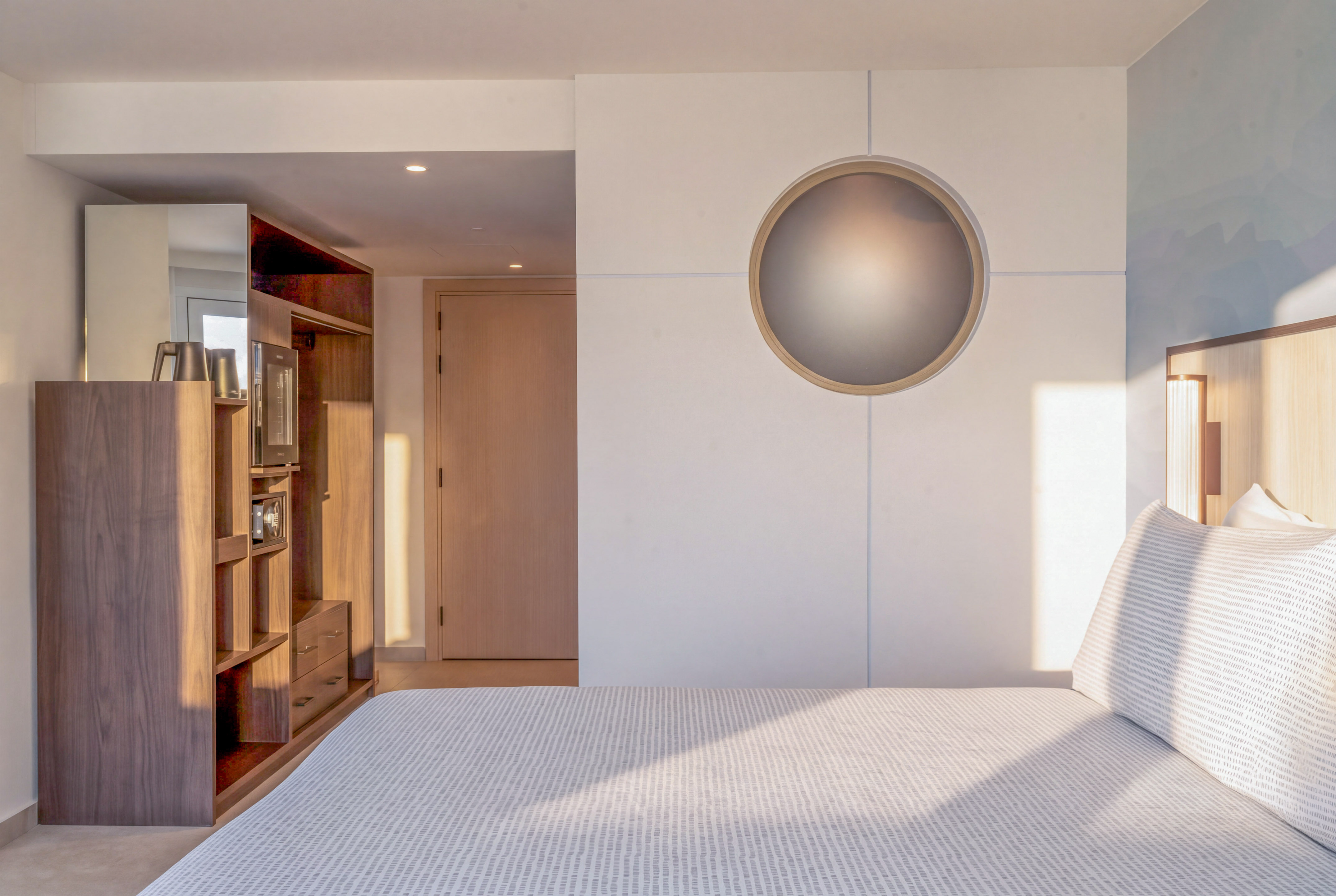 Modern hotel bedroom with bed, wooden storage, and circular wall light.