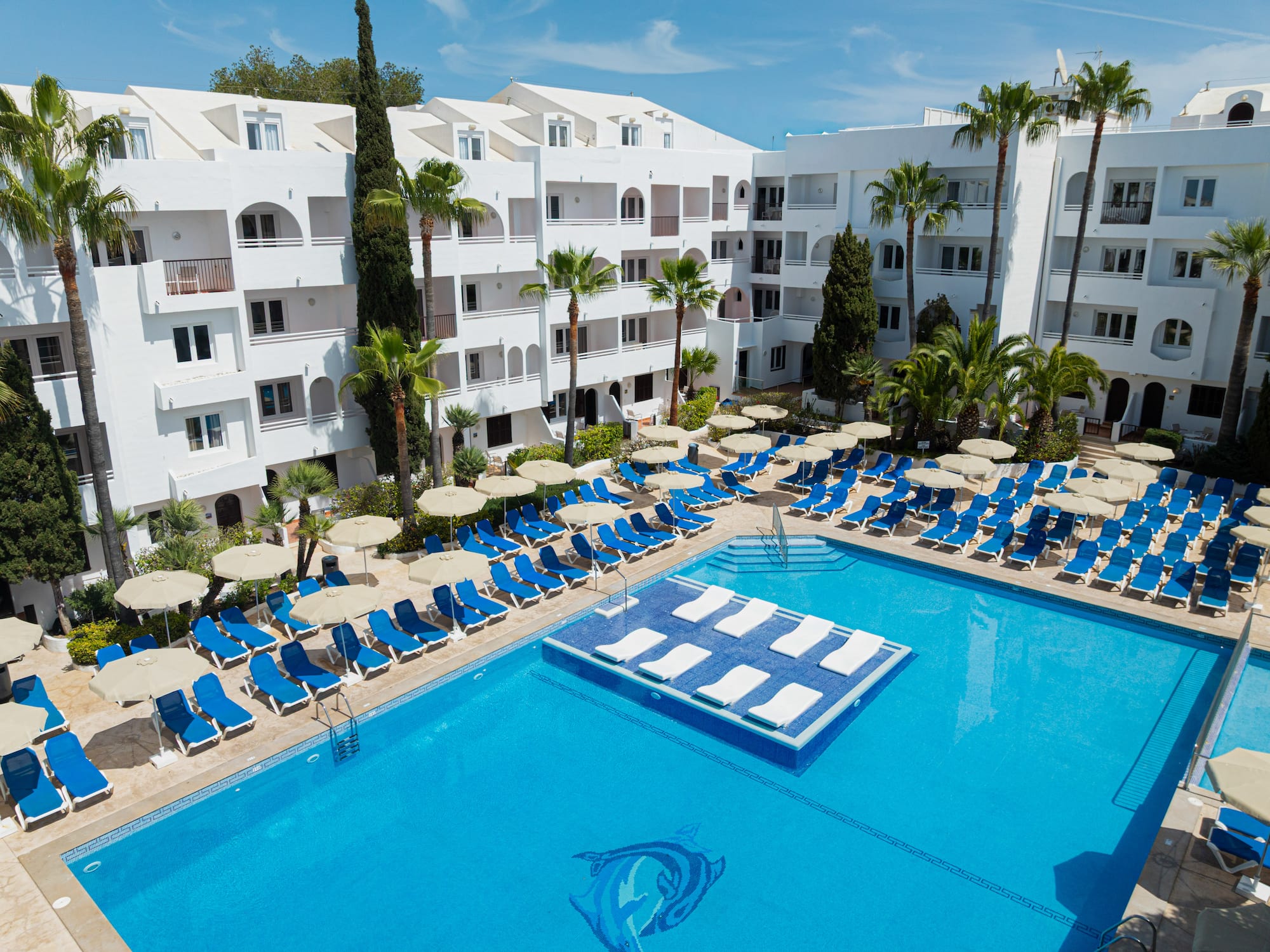 Sunny hotel pool with blue lounge chairs, palm trees, and white buildings.