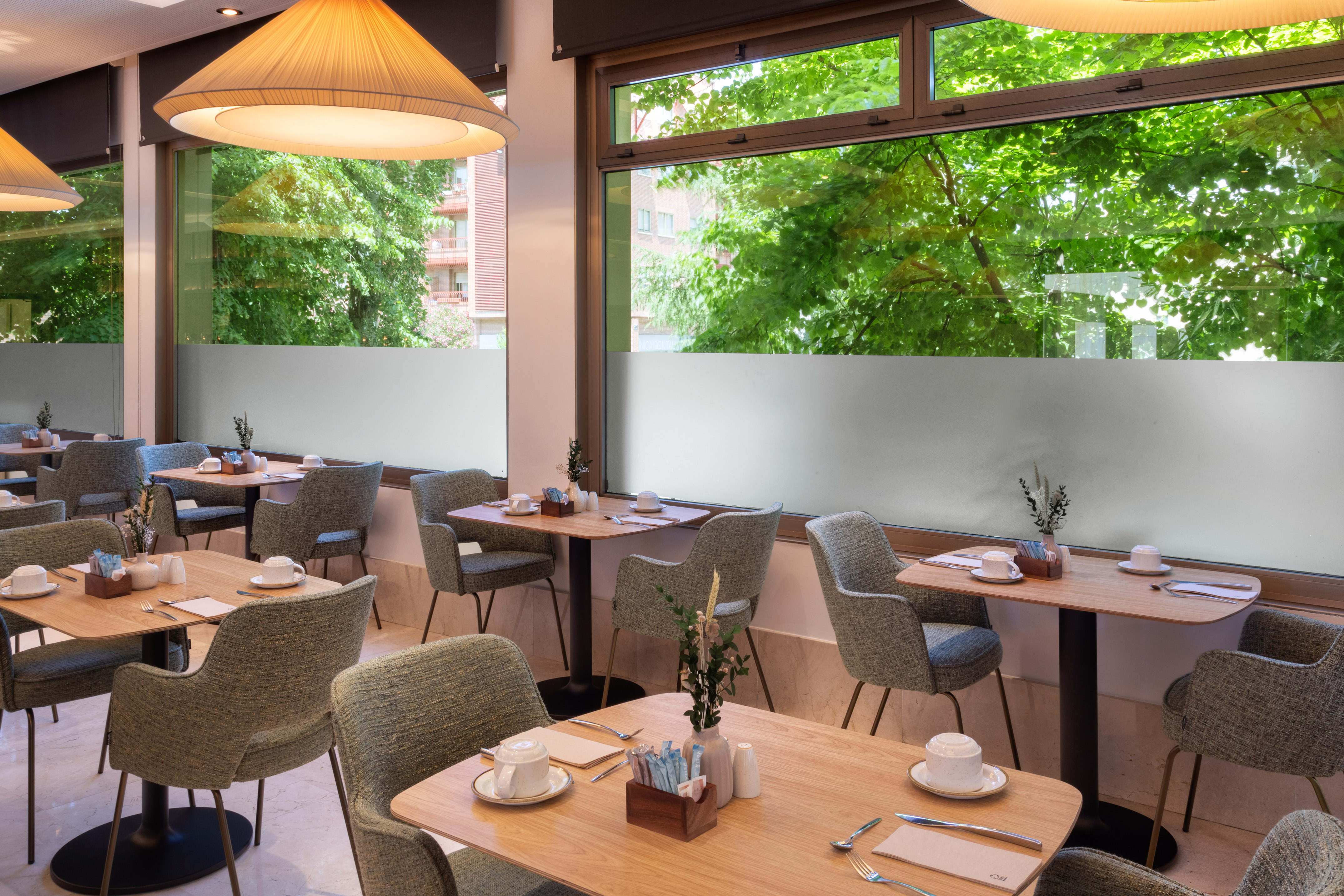 Modern restaurant with set tables, comfortable chairs, and green outdoor view.
