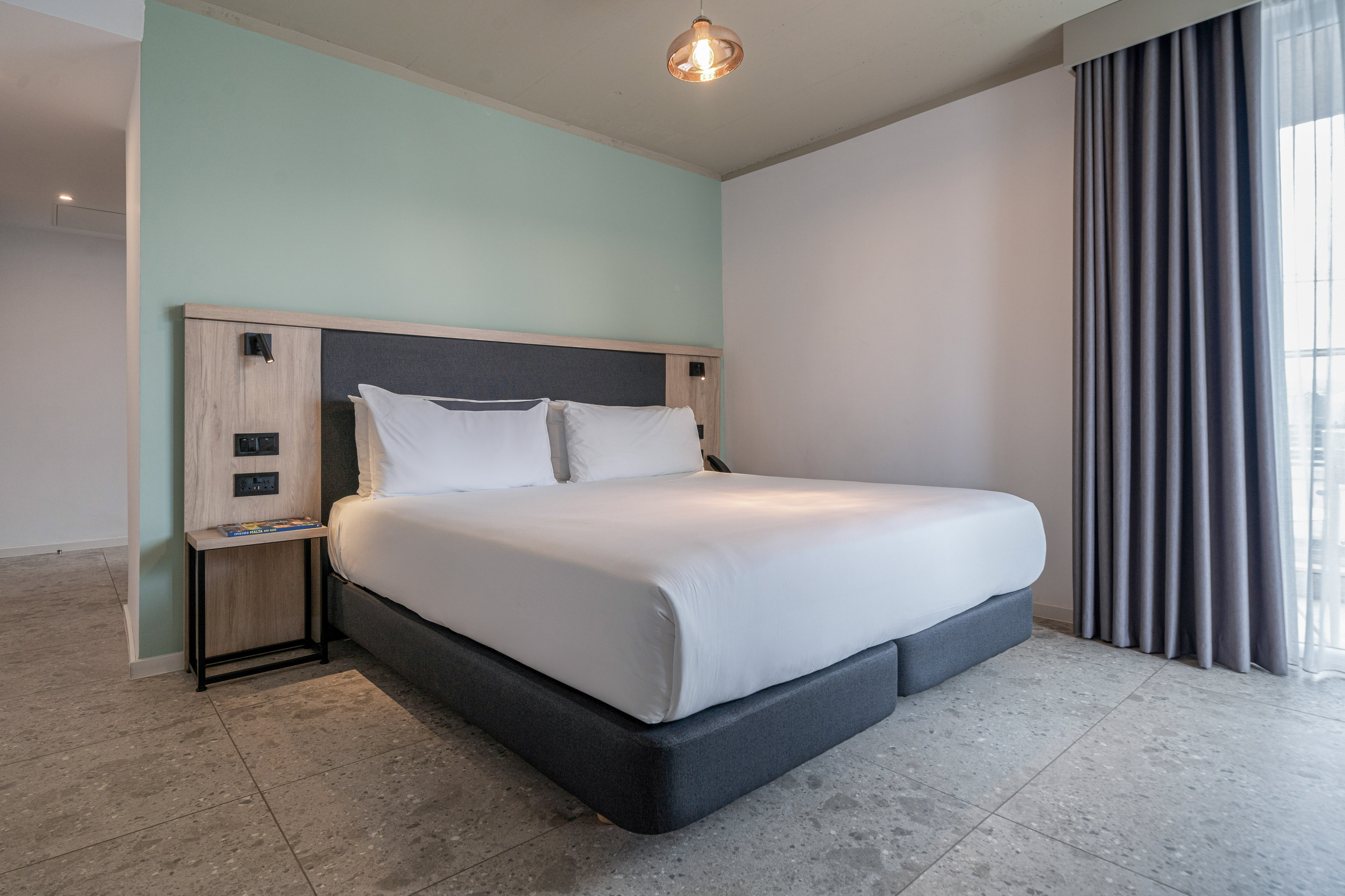 Modern hotel bedroom with white queen bed, mint accent wall, and gray curtains.