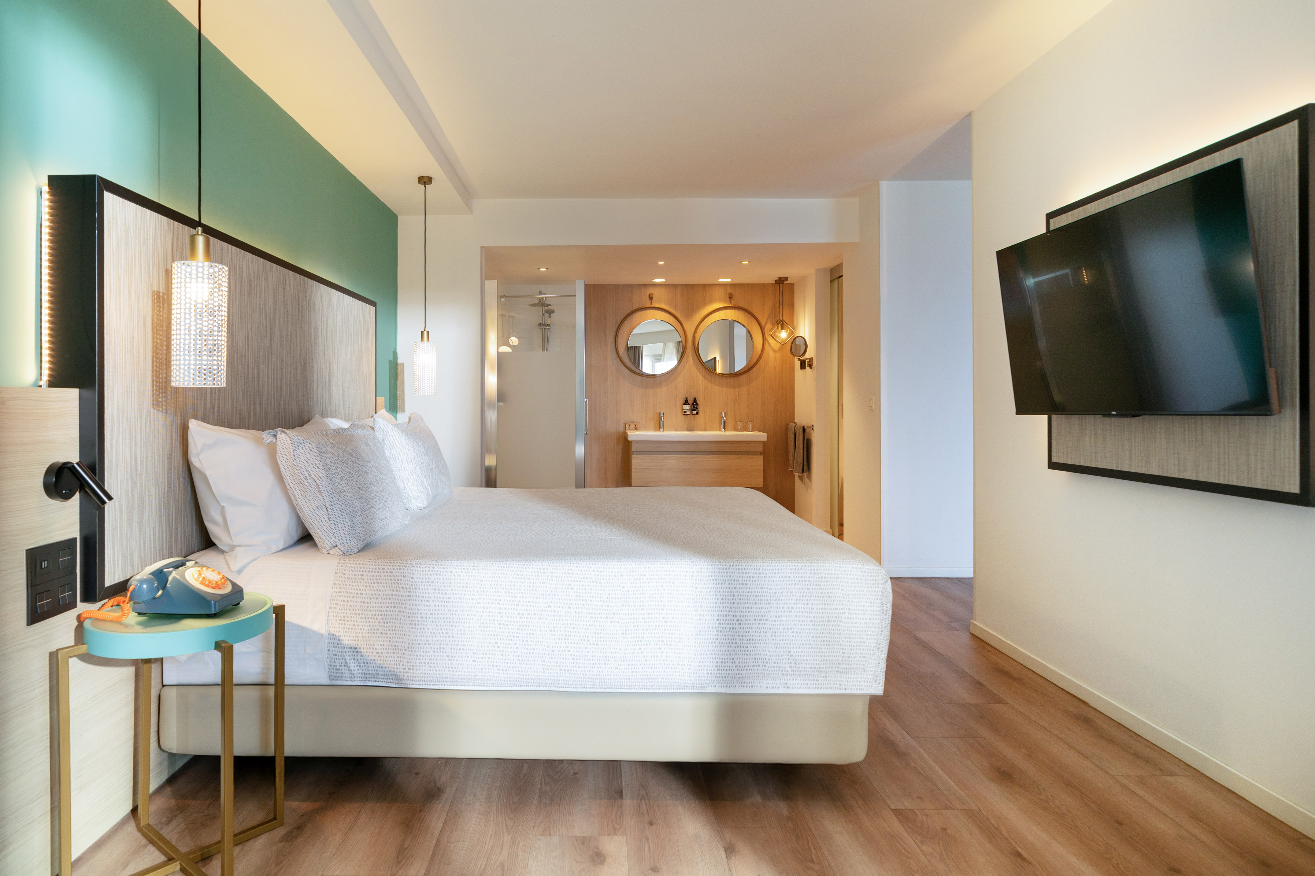 Modern hotel room with king bed, teal accent wall, wood floor, and open bathroom.