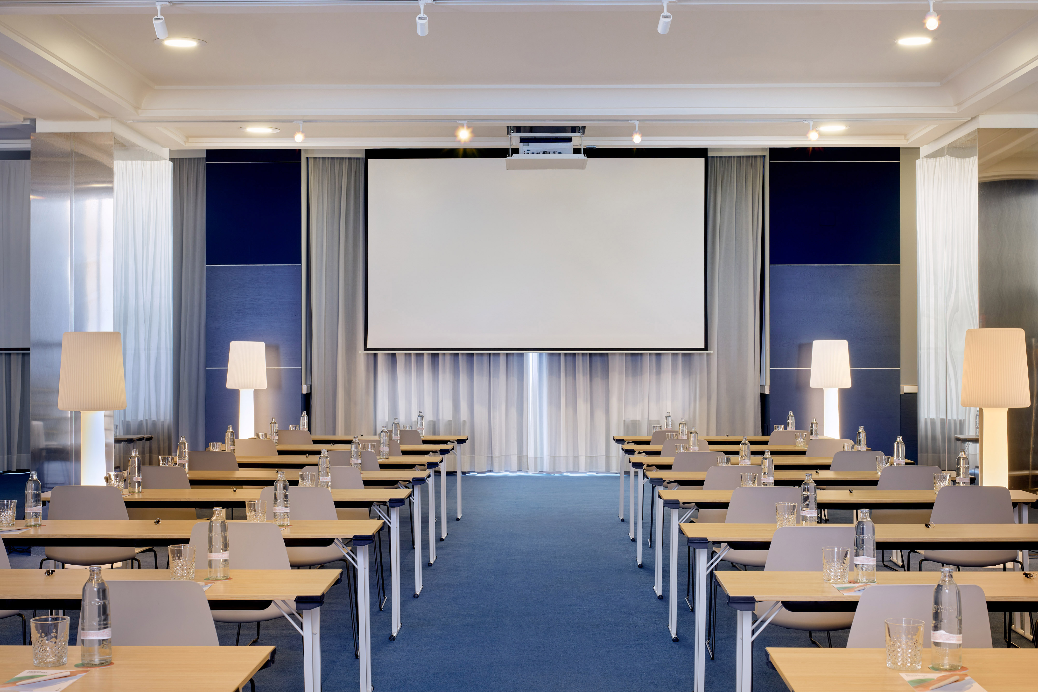 Sala de reuniones corporativa con pantalla, mesas, sillas y alfombra azul.