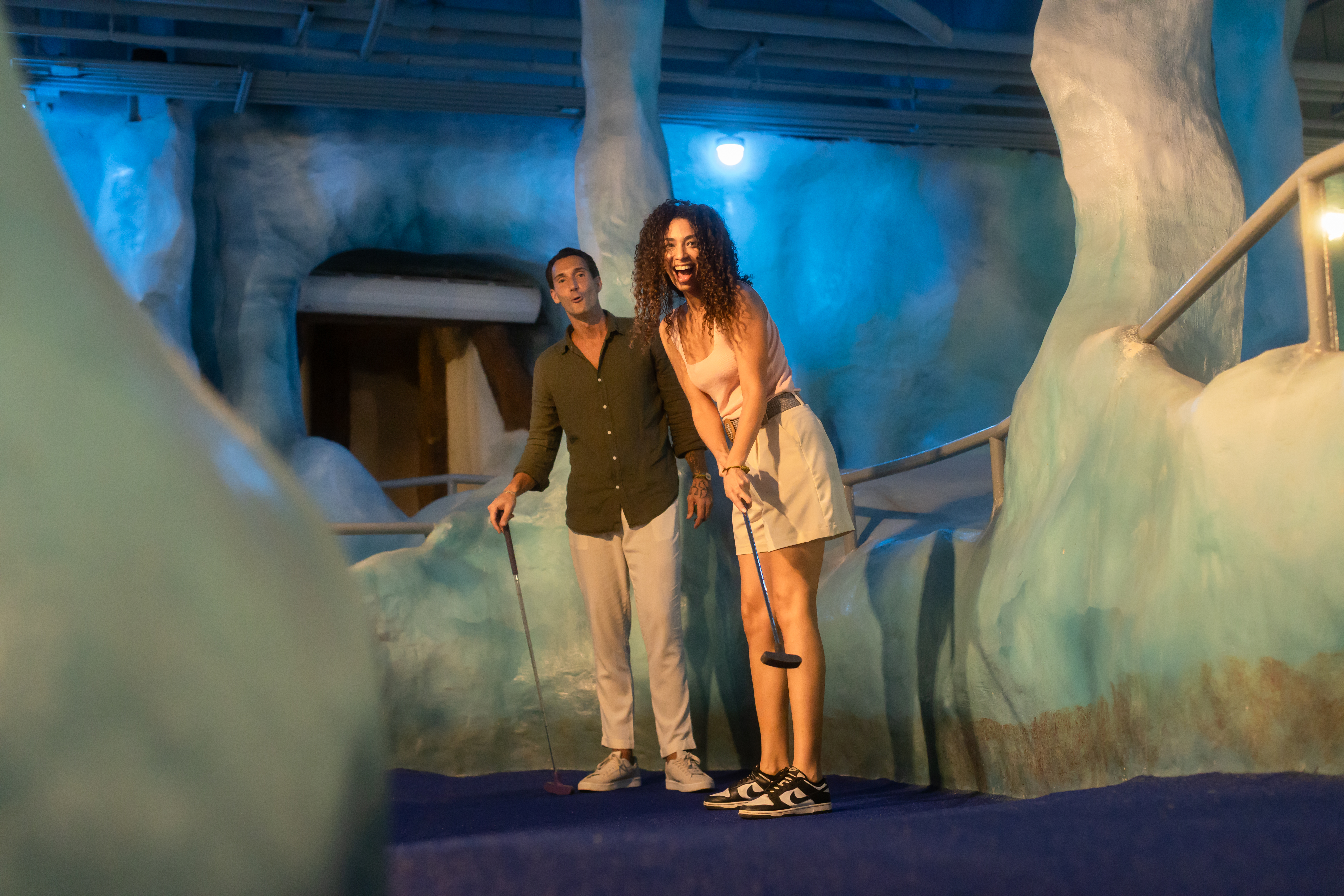 Happy couple enjoying indoor glow mini golf with putters in a themed cave.