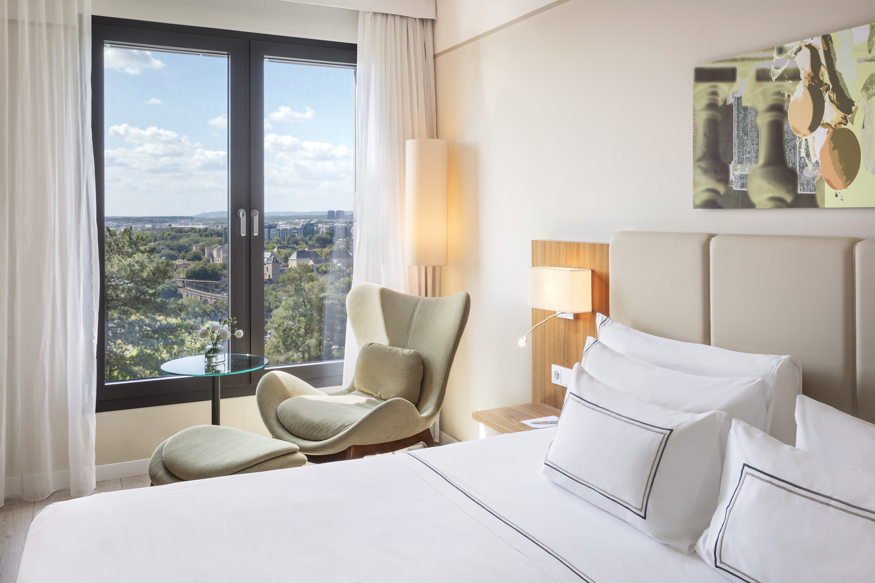 Luxury hotel room with city view, white bed, and comfortable armchair.