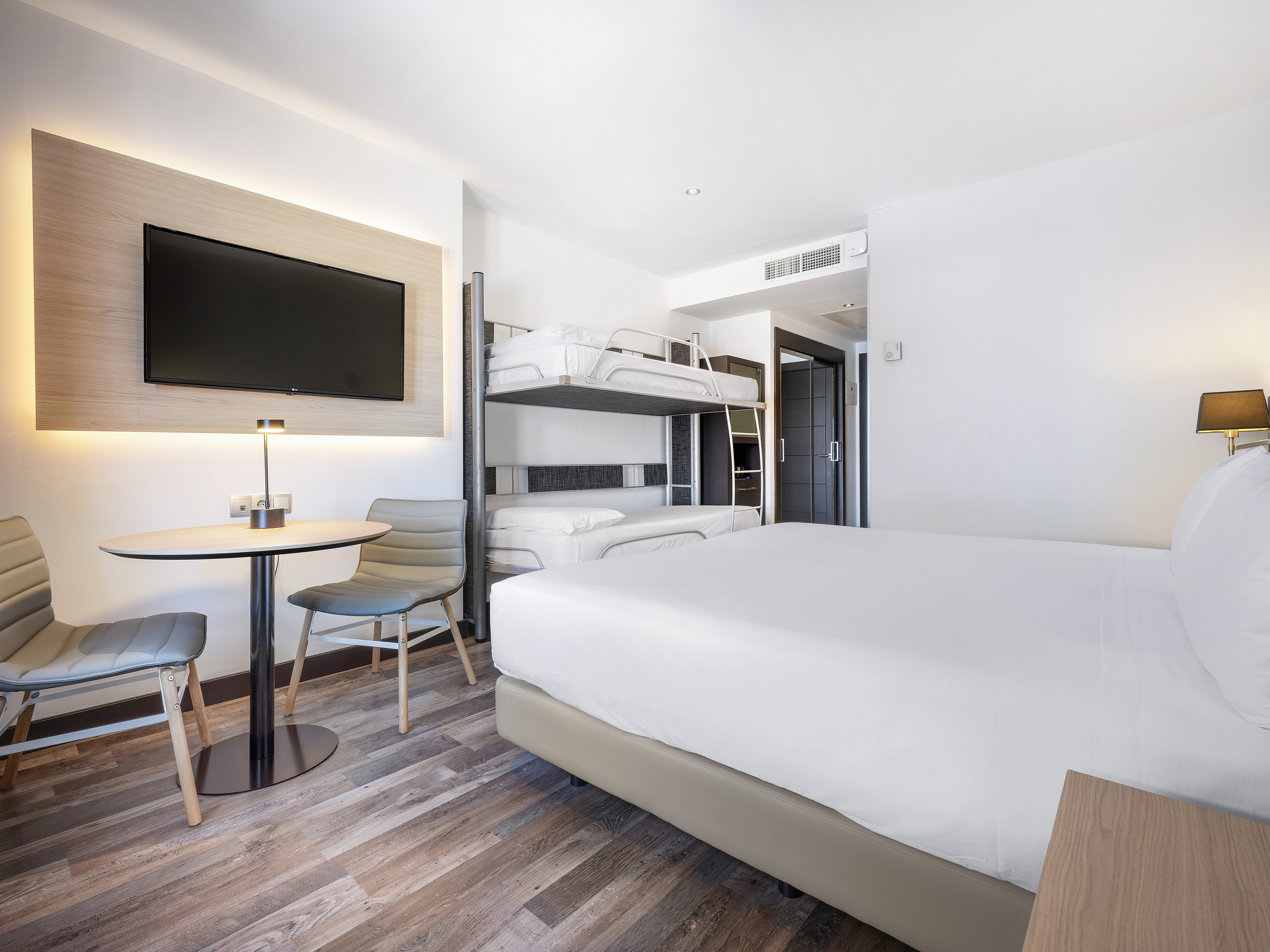 Modern hotel room with bed, bunk beds, wall-mounted TV, and small table.