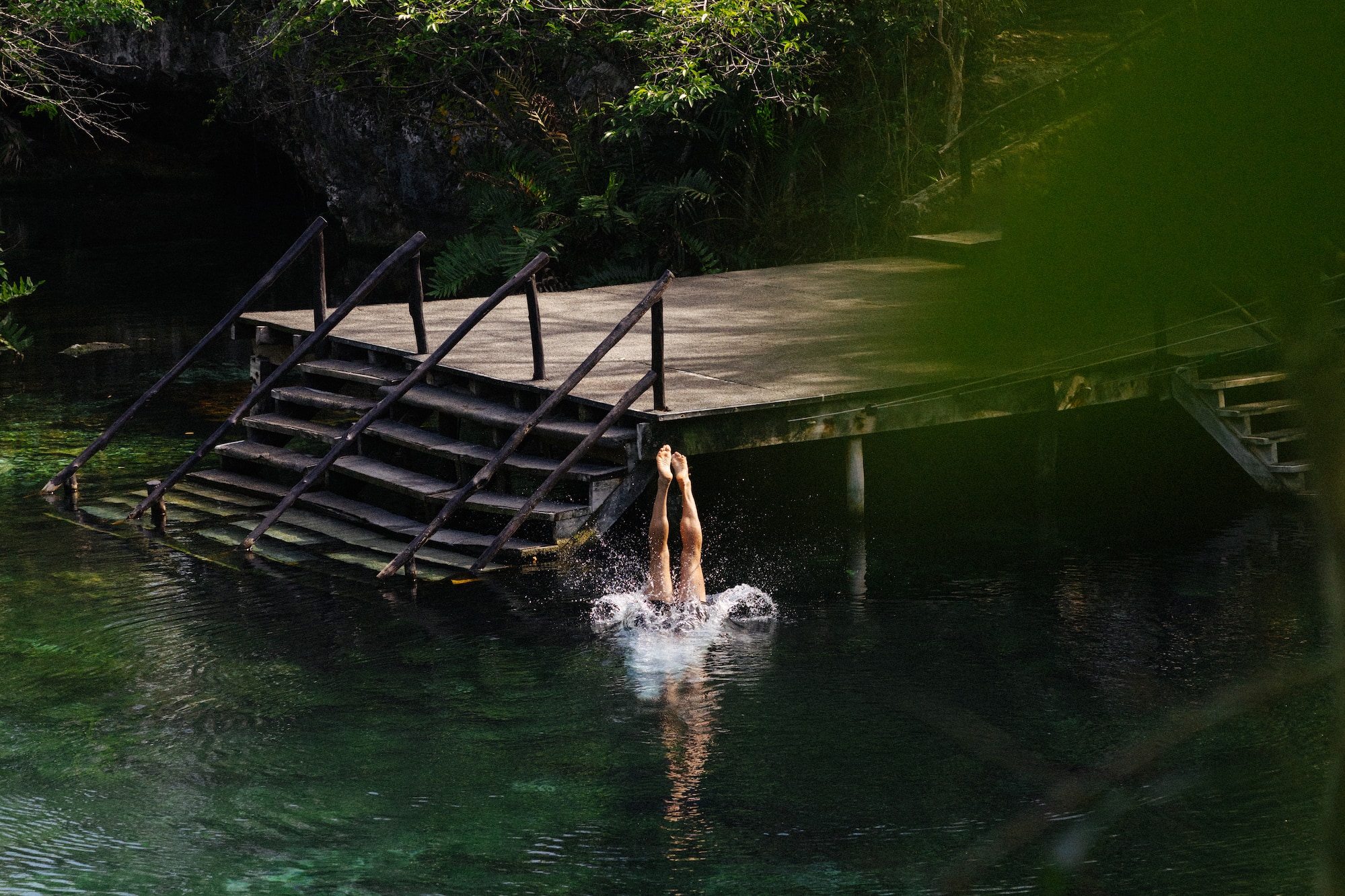 Diver plunging into clear green cenote water from wooden dock, lush jungle.