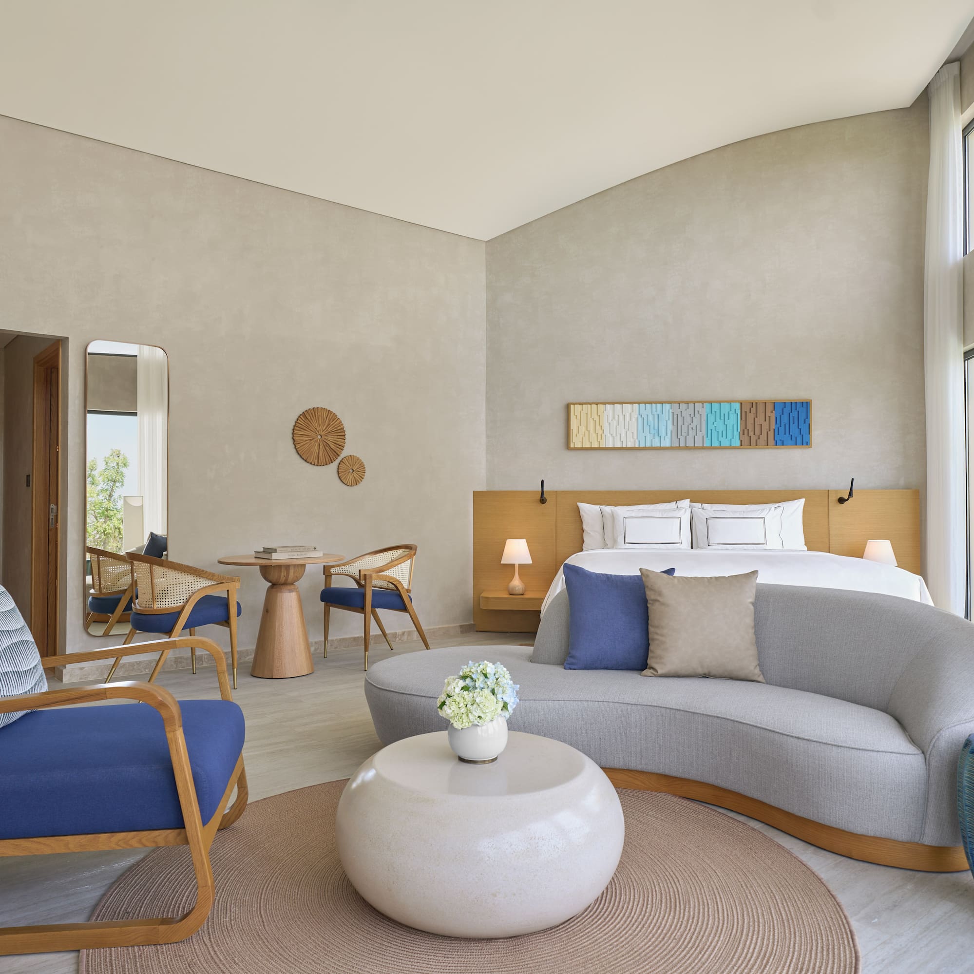 Modern hotel suite: bed, sofa, chairs, and large bright windows.