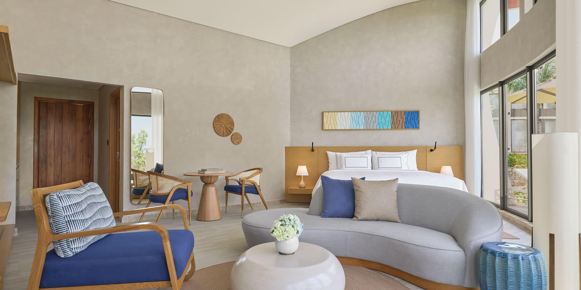 Modern hotel suite: bed, sofa, chairs, and large bright windows.
