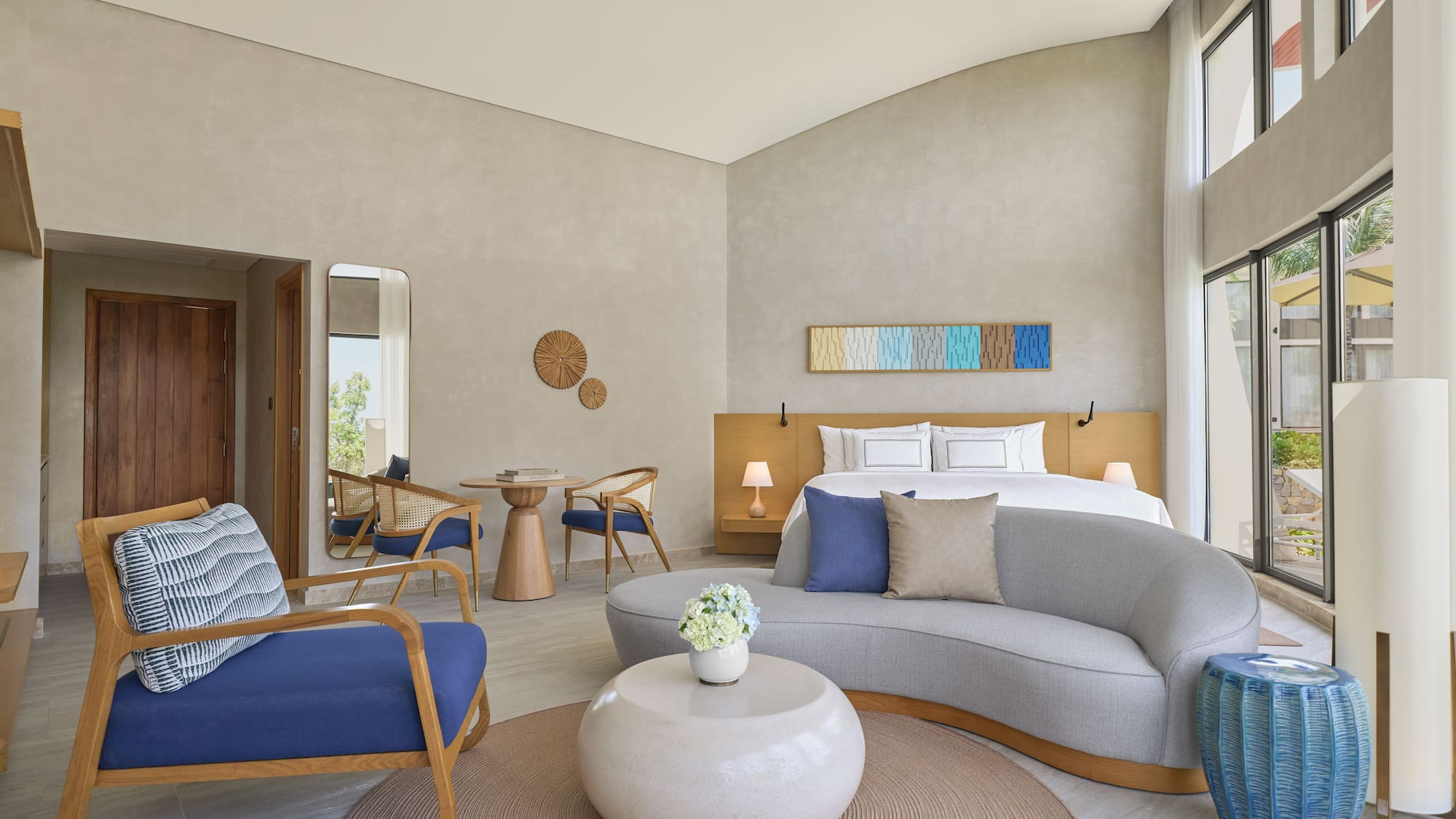 Modern hotel suite: bed, sofa, chairs, and large bright windows.