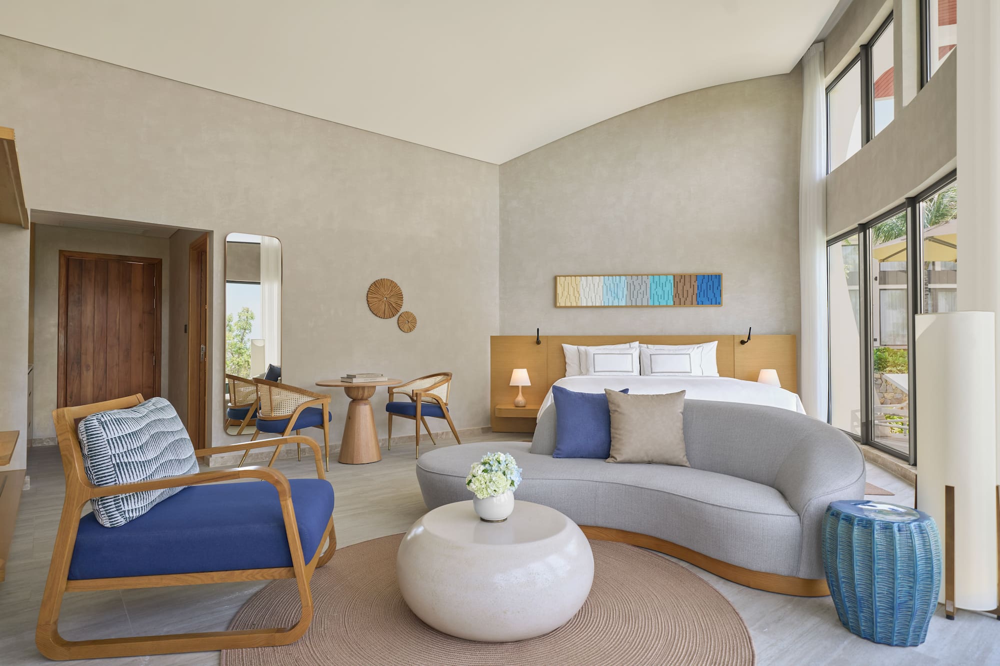 Modern hotel suite: bed, sofa, chairs, and large bright windows.