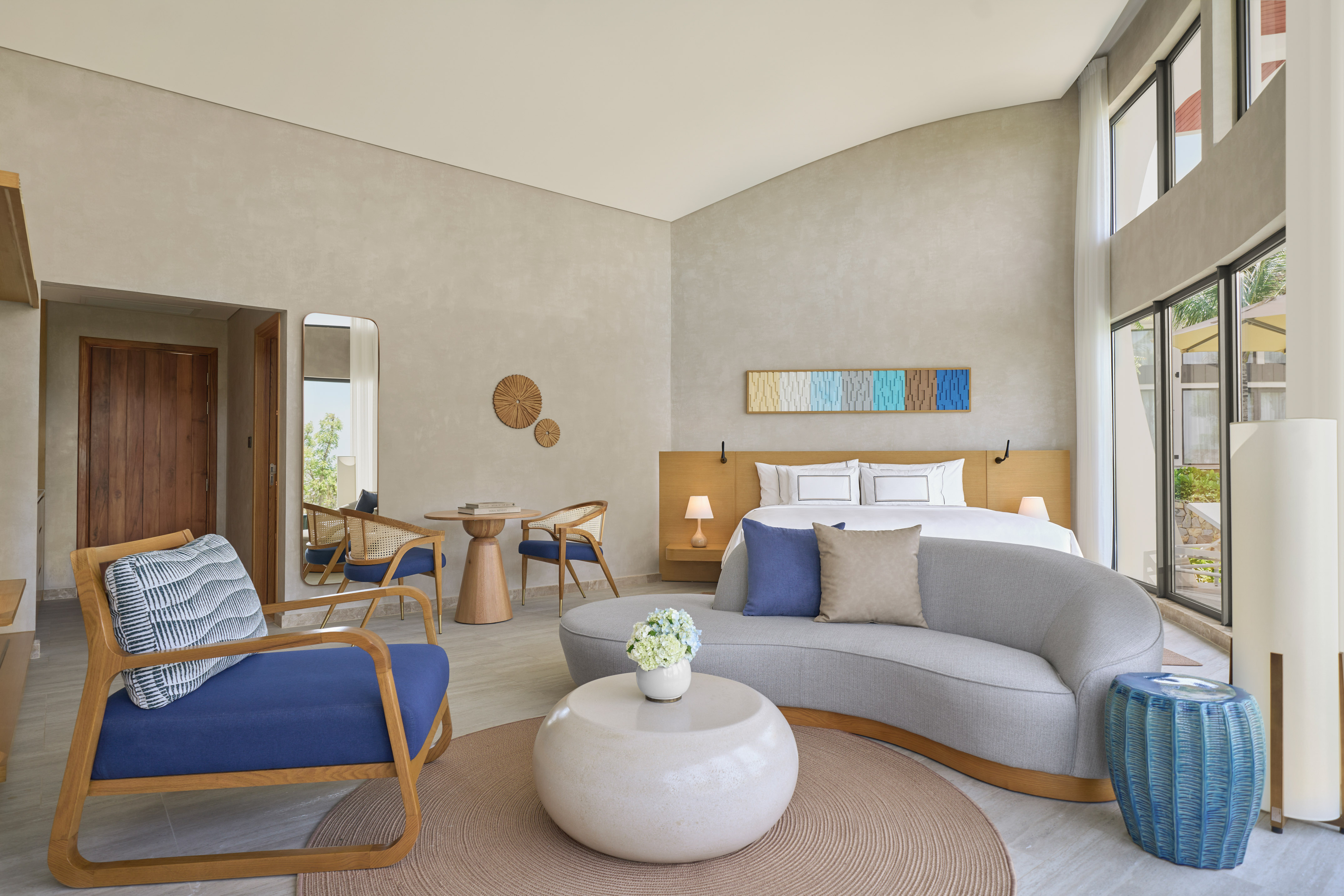 Modern hotel suite: bed, sofa, chairs, and large bright windows.
