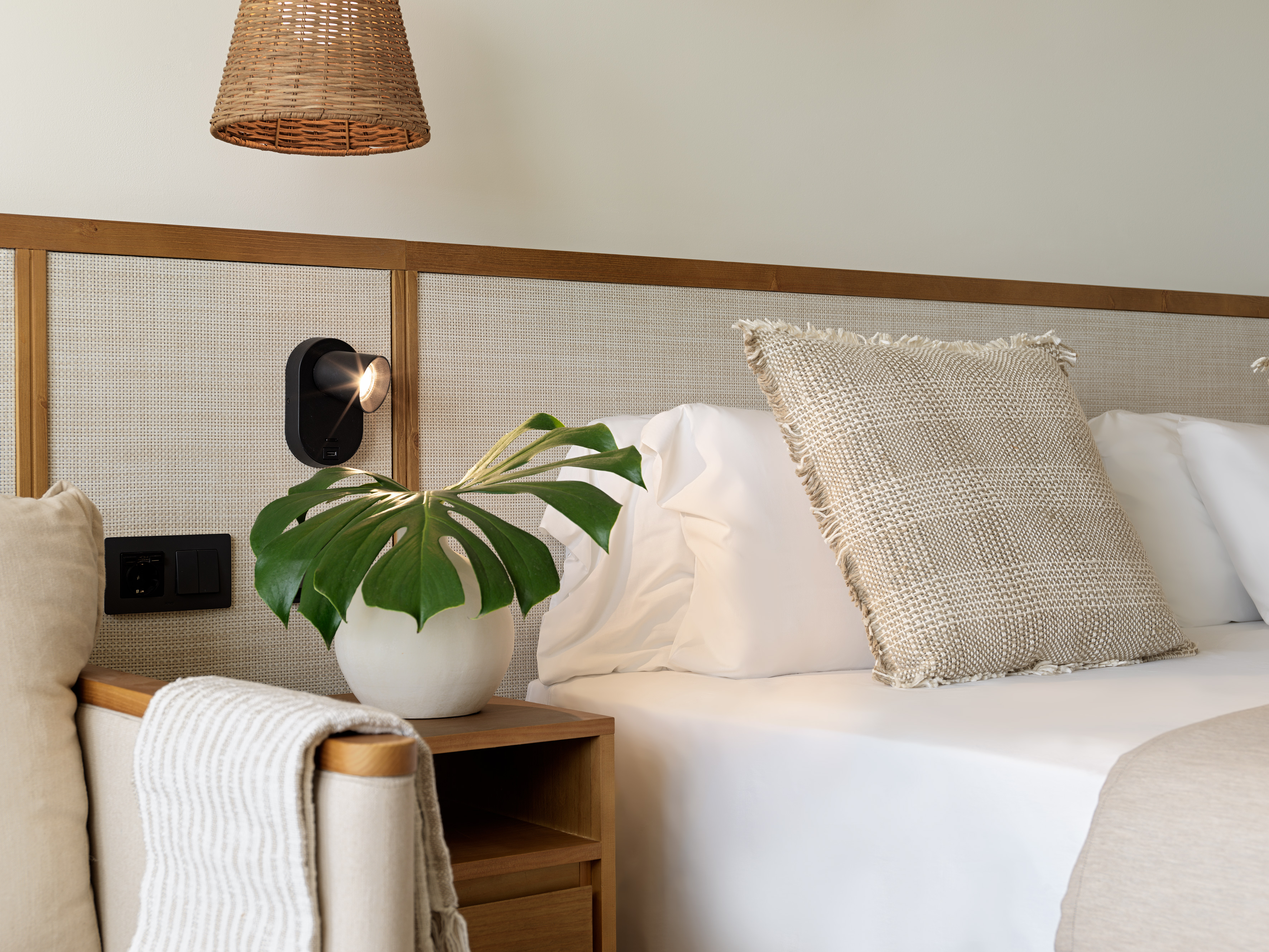Modern bedroom: white bed, cane headboard, wood nightstand, Monstera plant.