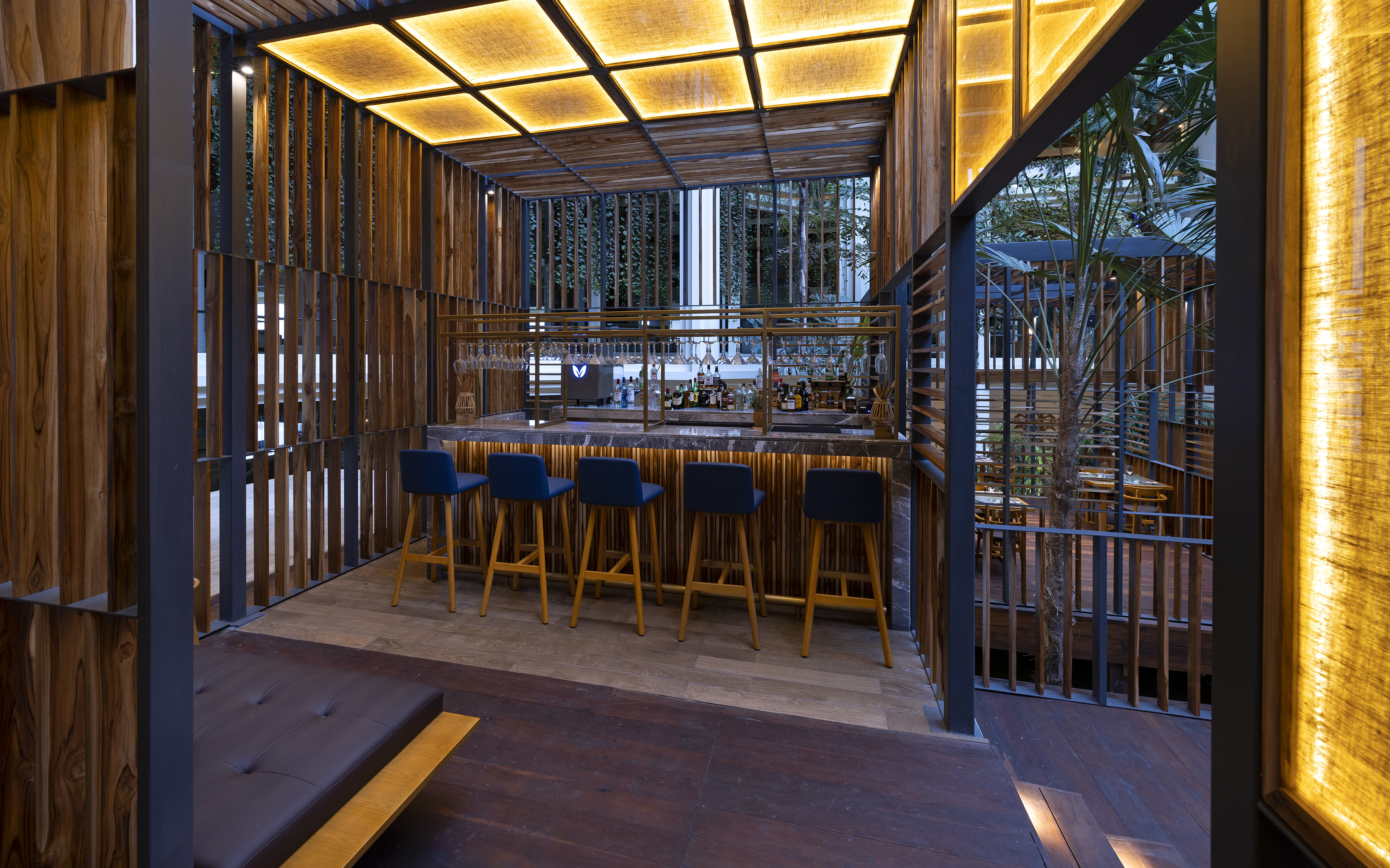 Modern outdoor bar with wood slatted design, warm lighting, blue stools.