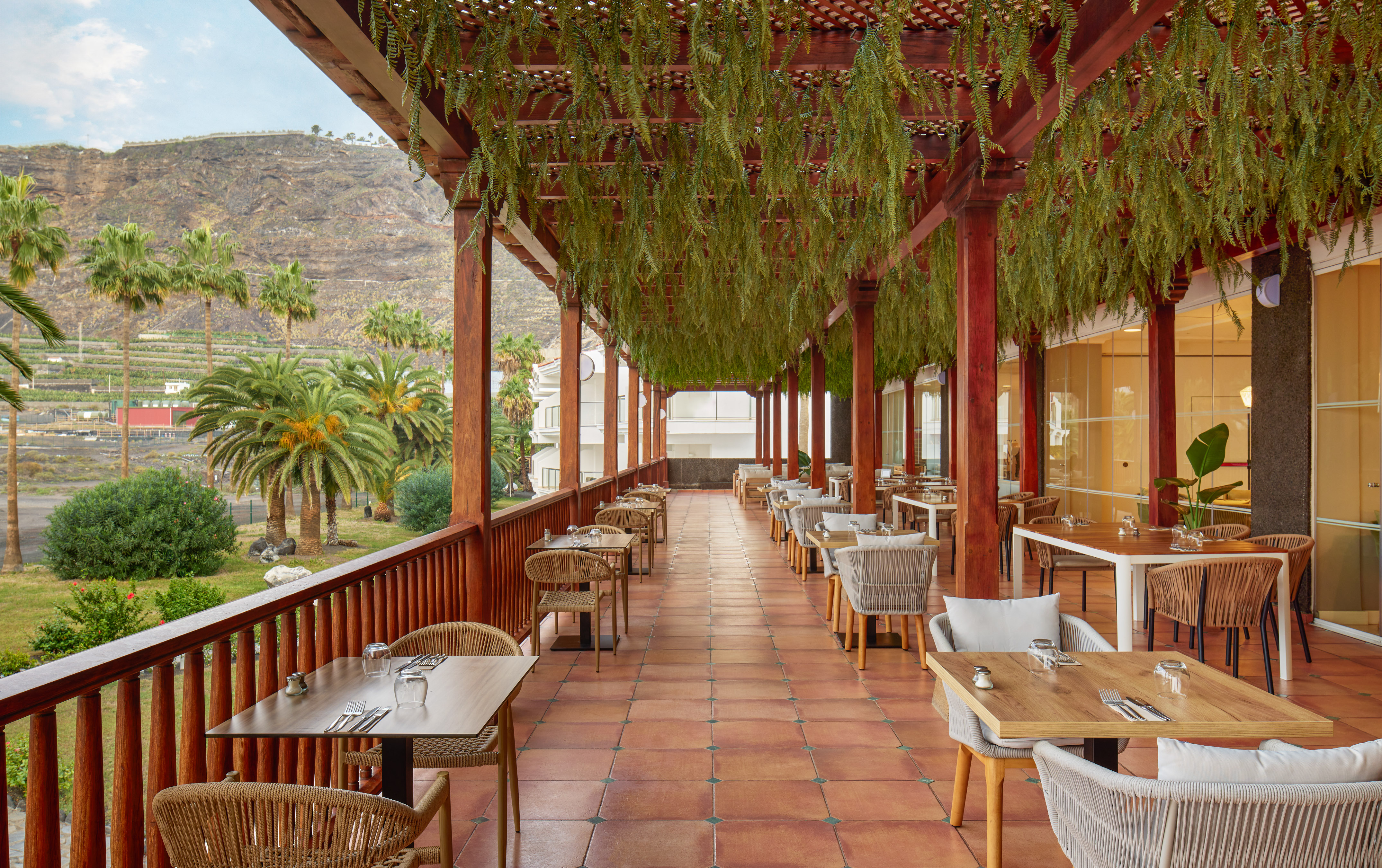 Outdoor restaurant terrace with green pergola, palm trees, and mountain backdrop.